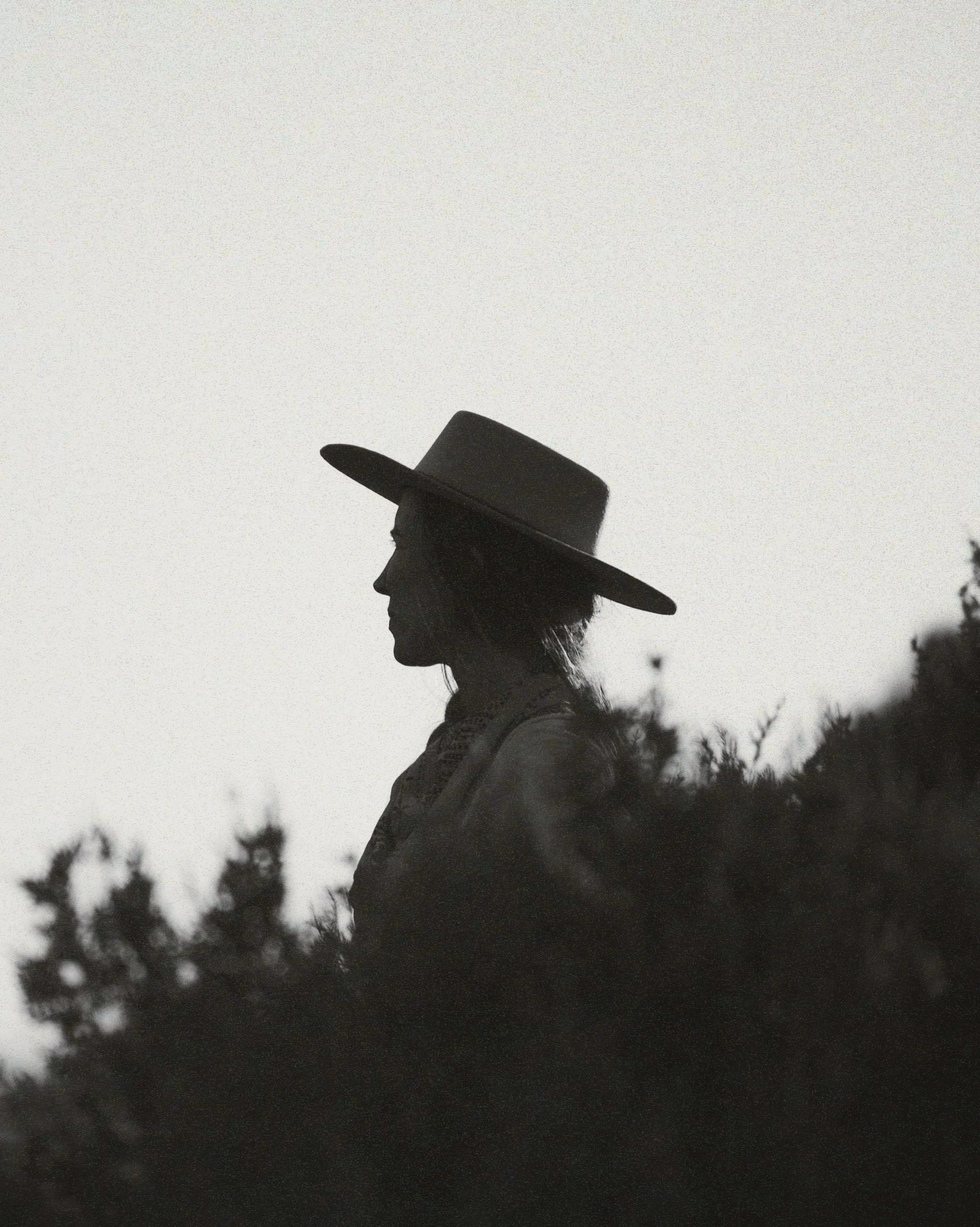 Silhouette of a woman wearing a wide-brimmed hat, facing left, with foliage in the foreground and sky in the background.