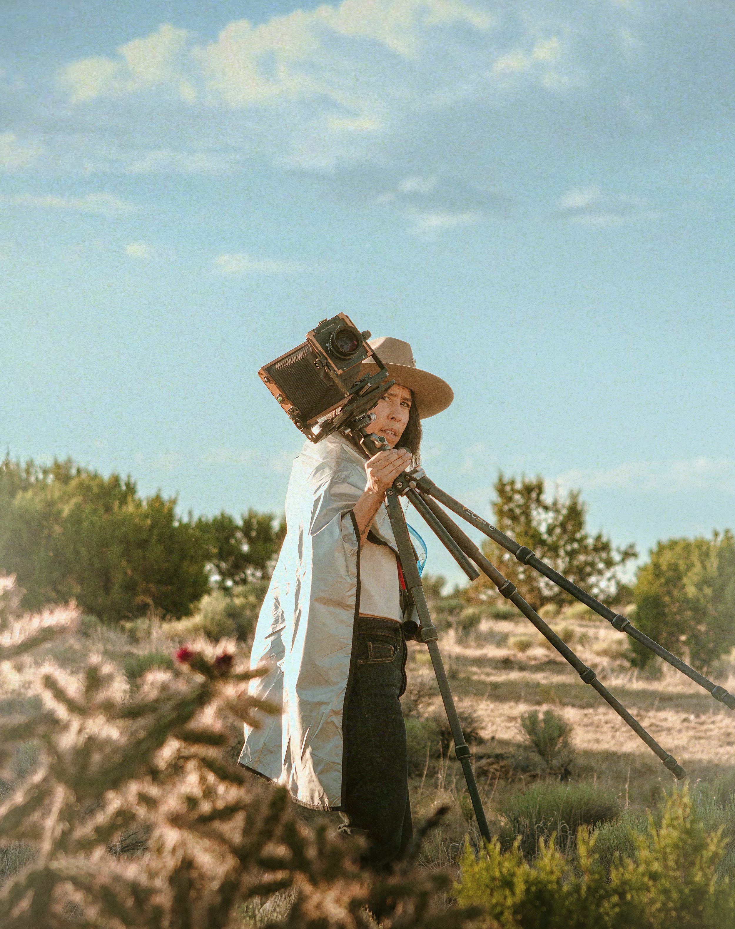 A woman wearing a wide-brimmed hat stands outdoors, operating a professional camera mounted on a tripod.