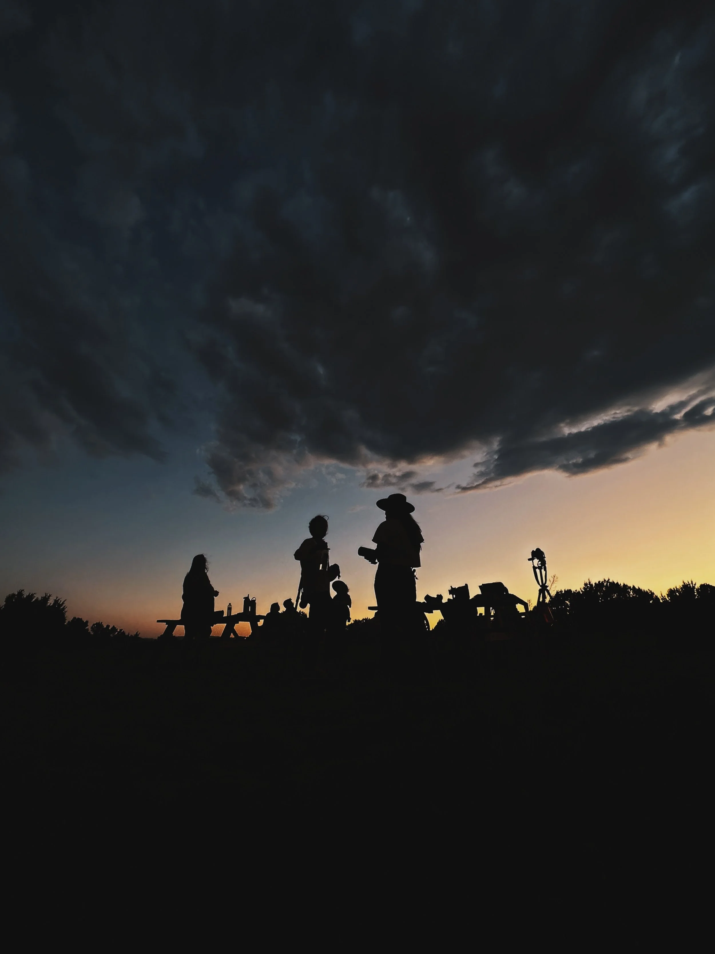Silhouettes of people gathered outdoors at sunset, with a dark, cloudy sky overhead.
