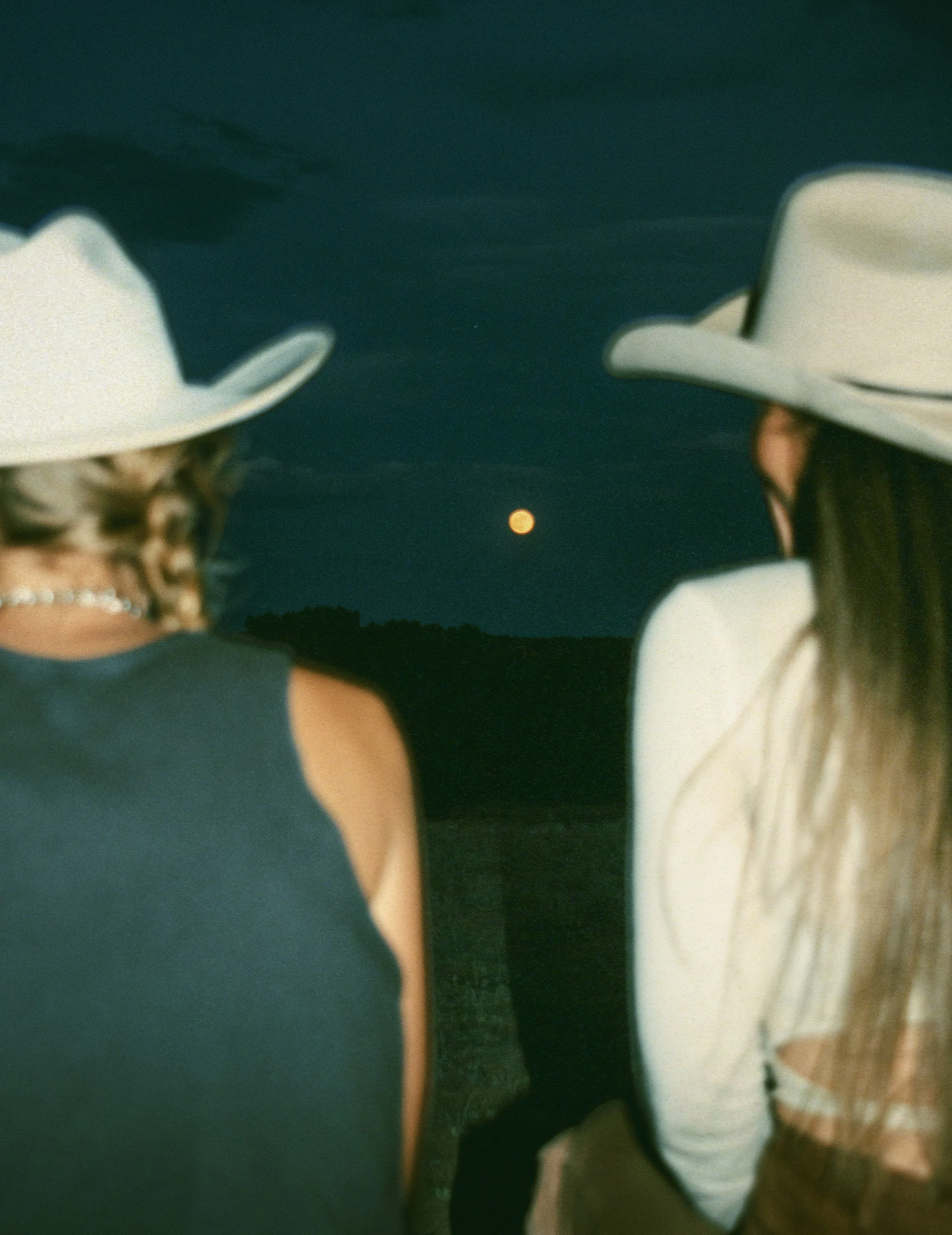 Two women wearing white cowboy hats standing outdoors at night, looking at a bright moon in the sky.