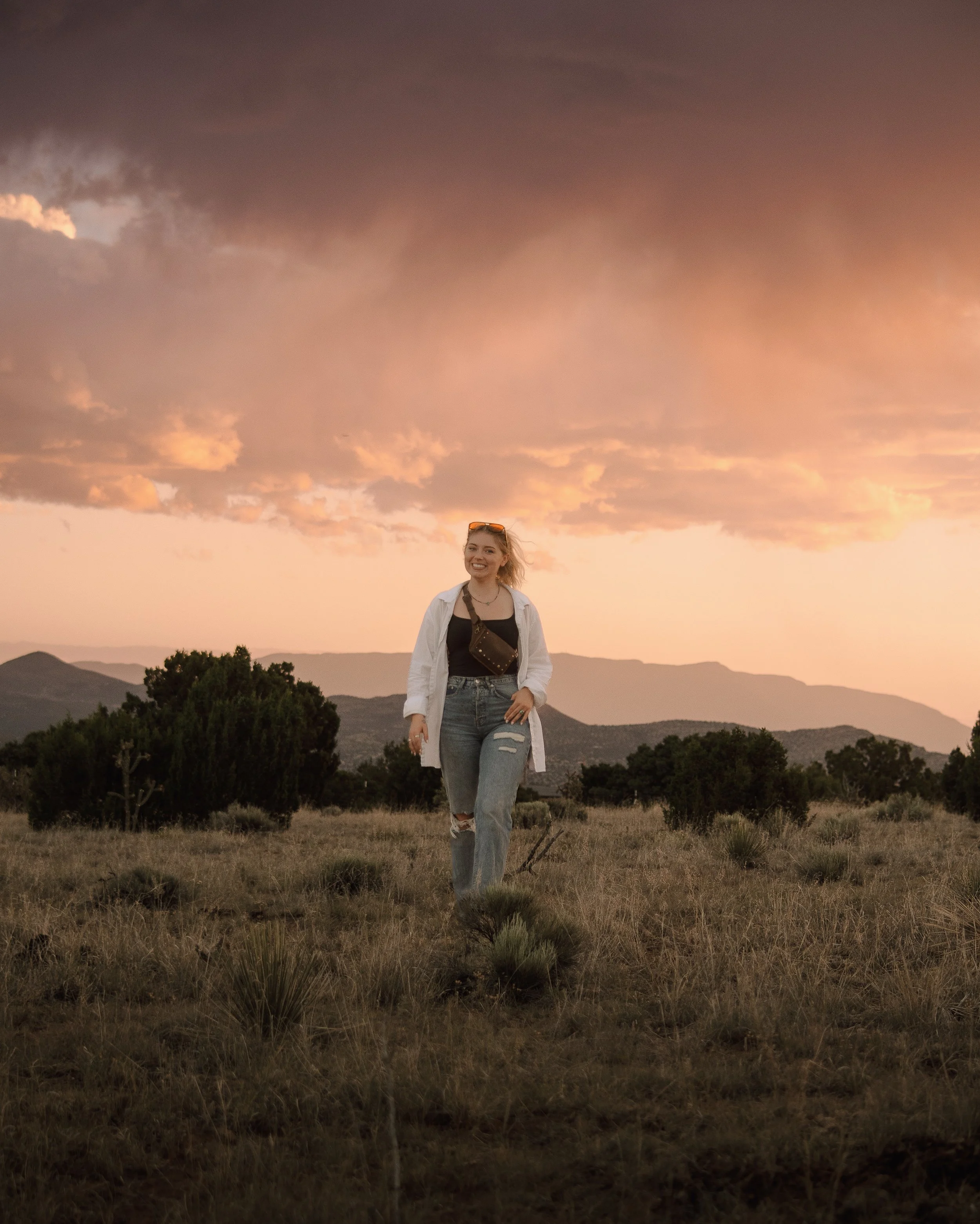 A young woman smiling and walking in a grassy field during sunset, with mountains and trees in the background.