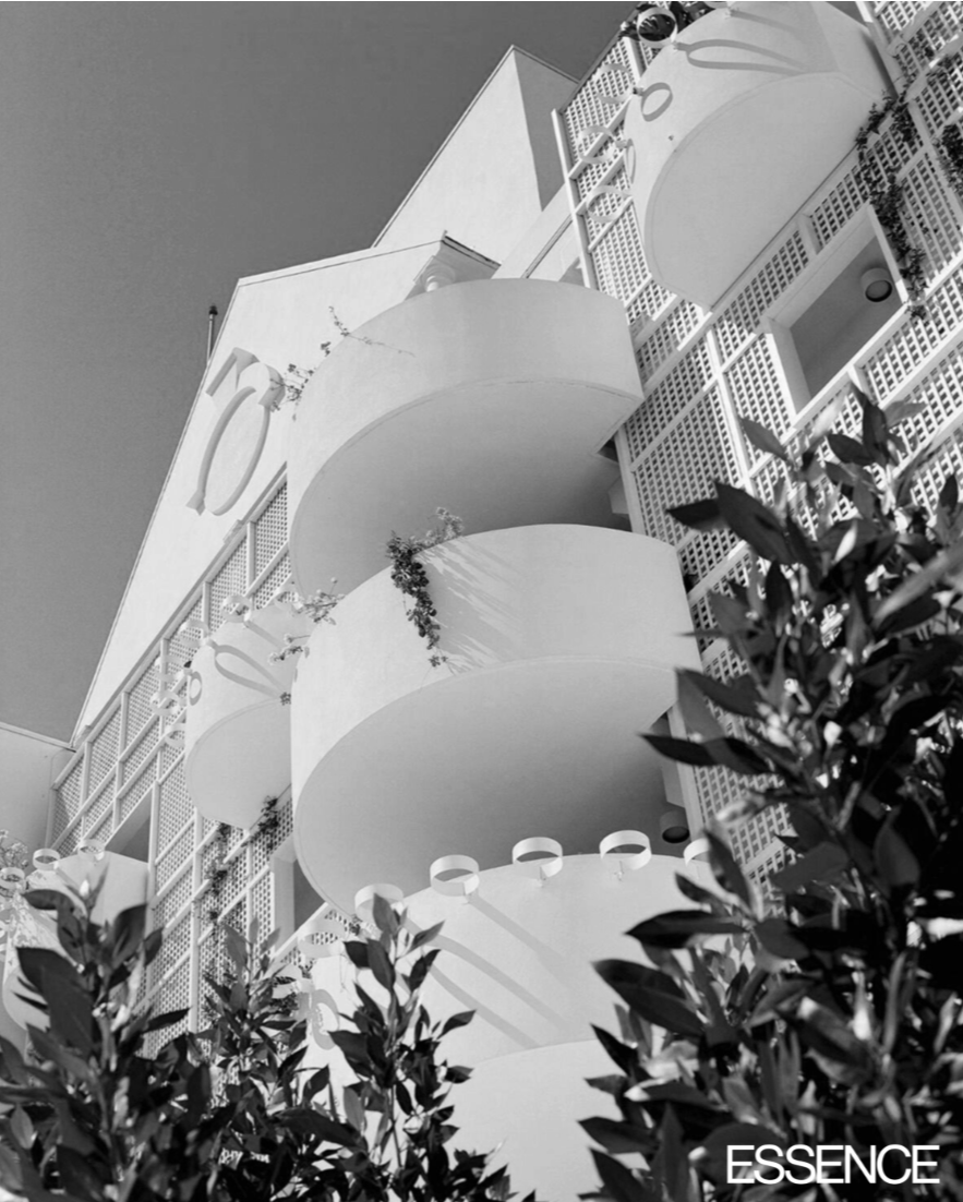 A black-and-white photo of a modern building with curved balconies and lattice design, partially obscured by foliage in the foreground. The building has the number '10' visible on its facade, and the word 'ESSENCE' is written in the bottom right corner.