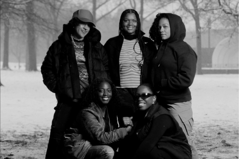 Five women posing outdoors on a snowy day, with trees and a building in the background, dressed in winter clothing.