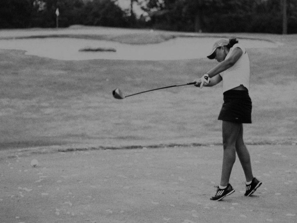 A woman playing golf on a golf course, preparing to hit a golf ball with a club, with trees and a golf green in the background.