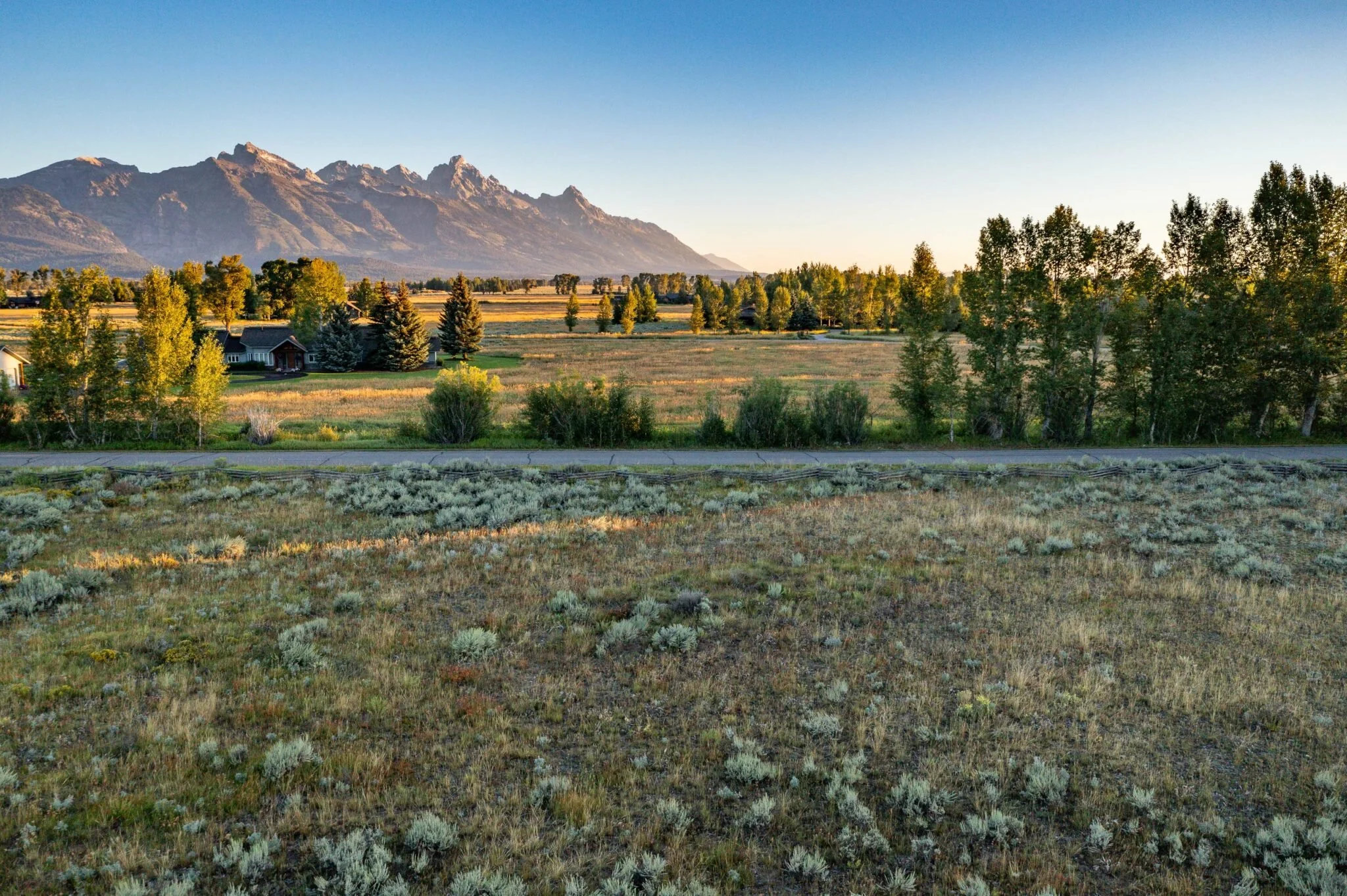 Scenic landscape with mountains in the background, trees, a few houses, open fields, and a dirt path in the foreground, illuminated by sunlight.