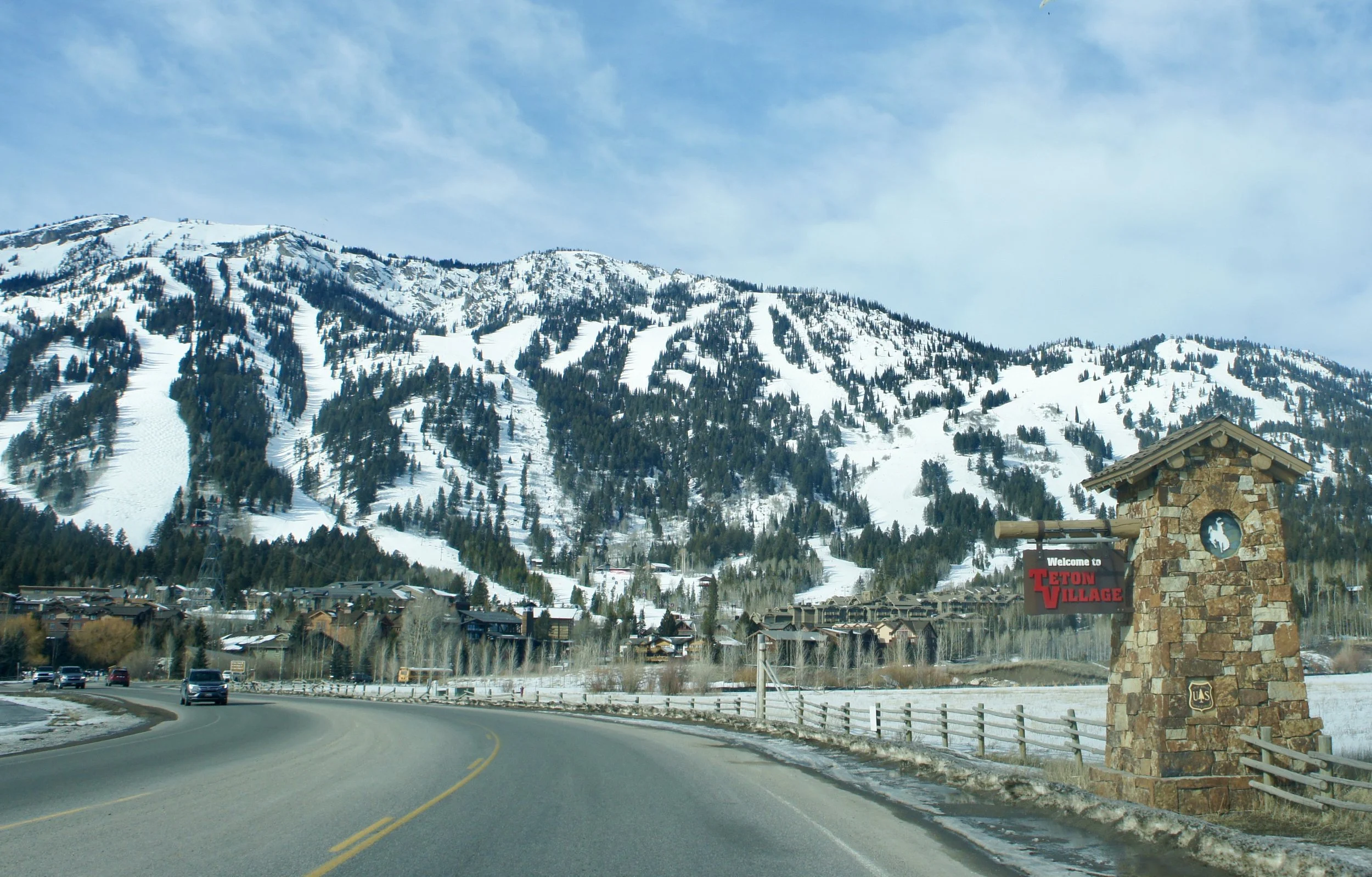 View of a mountain with snow and trees, a winding road, and a stone entrance sign reading 'Welcome to Teton Village' in Wyoming.