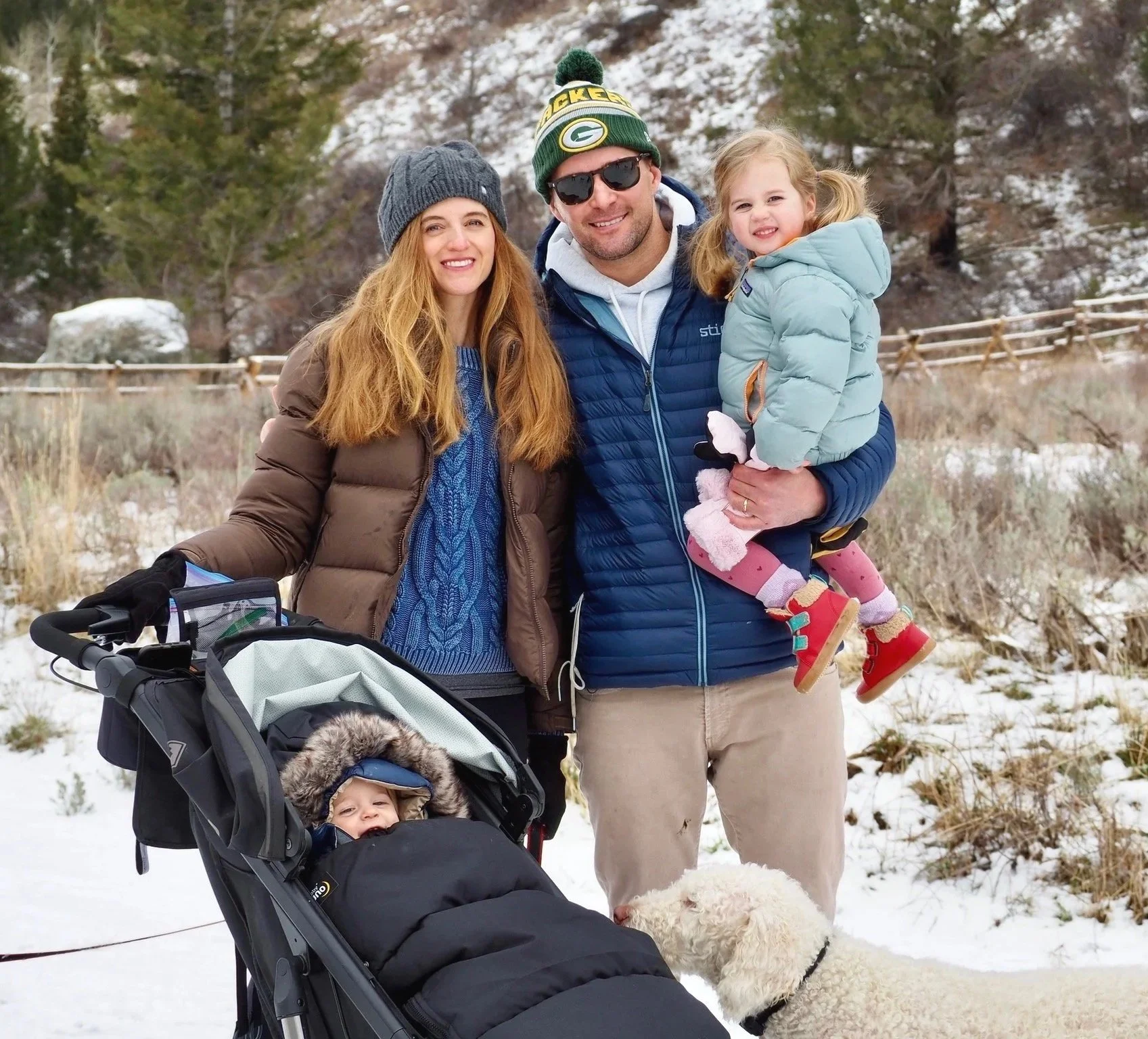 A family of four, with two young children, is enjoying a snowy outdoor scene. The mother, with long red hair and a gray beanie, stands next to a stroller with a baby inside, bundled up for winter. The father, wearing sunglasses, a green and yellow Green Bay Packers hat, and a blue jacket, is holding a young girl with pigtails in a light blue puffy jacket. A white dog is sniffing the baby, who is in the stroller.