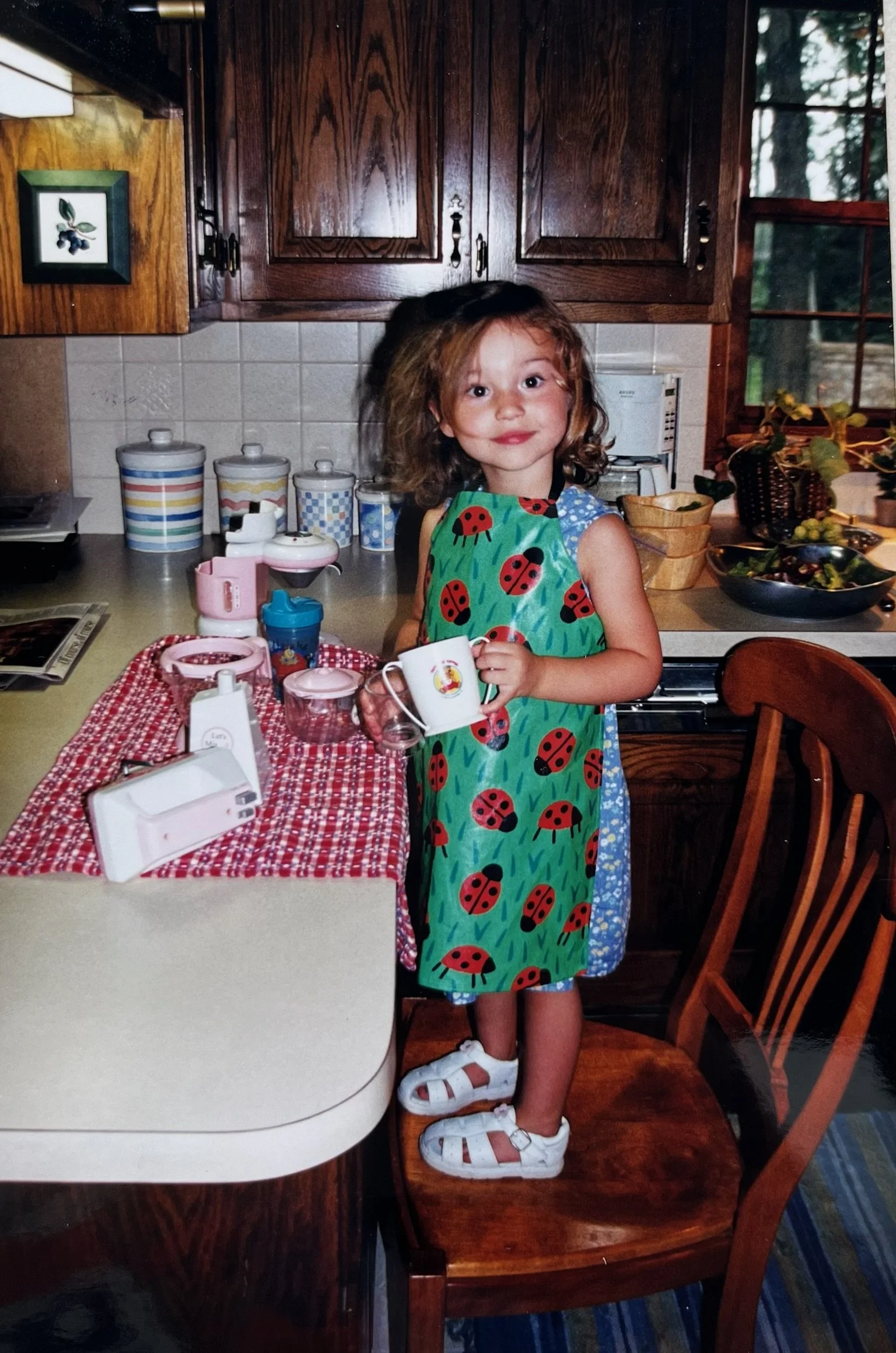 A young girl with curly hair, dressed in a ladybug-themed apron, holding a cup, standing on a wooden chair in a kitchen with wooden cabinets, a window showing trees outside, and a table with various dishes and kitchen items.