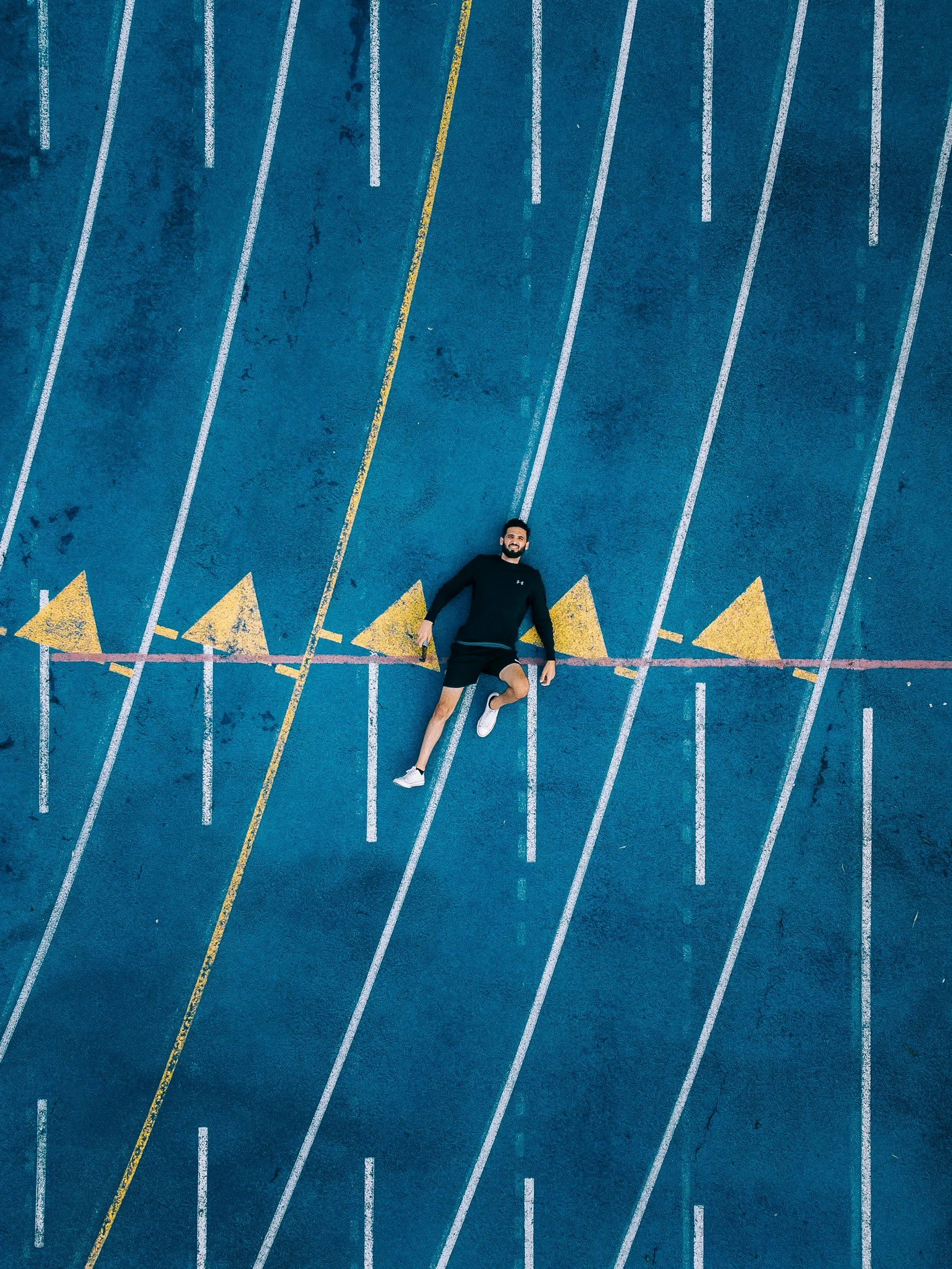 An overhead view of a man lying on a running track, smiling, surrounded by white lines and yellow triangle markings.