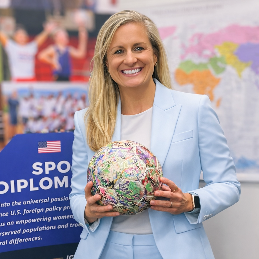 A woman in a light blue suit holding a colorful, painted soccer ball in a classroom with a world map and a blue sports diplomacy display in the background.