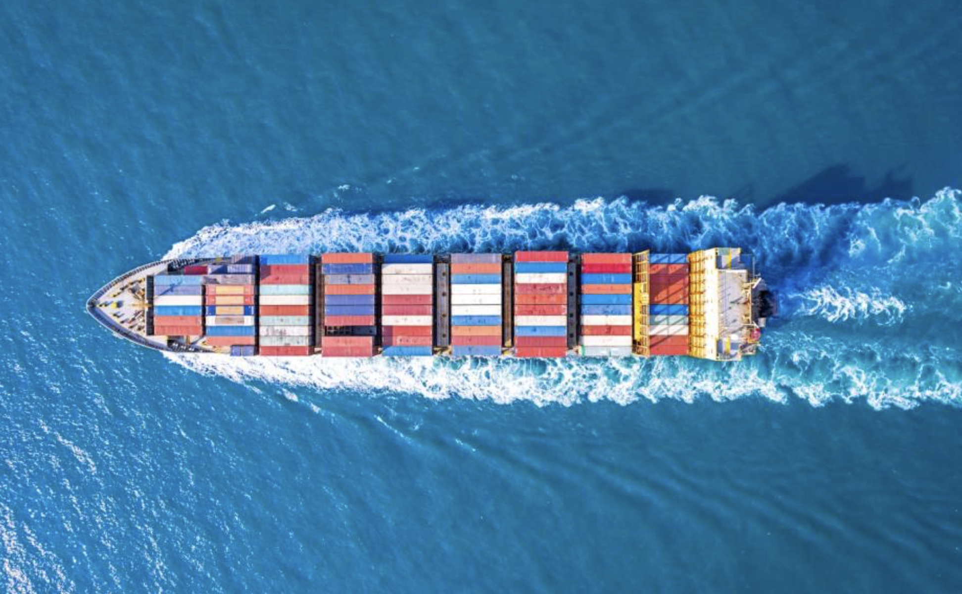 Bird's eye view of a cargo ship sailing through the ocean, carrying numerous multicolored shipping containers.