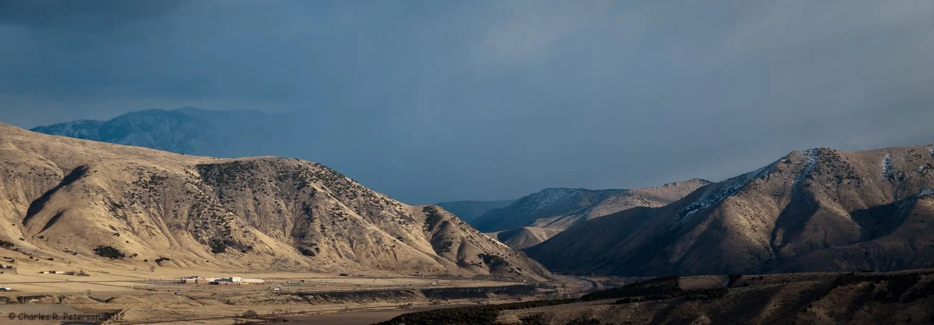 A panoramic view of dry, mountainous terrain with steep, rugged hills and snow-capped peaks in the distance. Sparse vegetation is visible on the hills, and a few buildings are present at the base of the mountains.