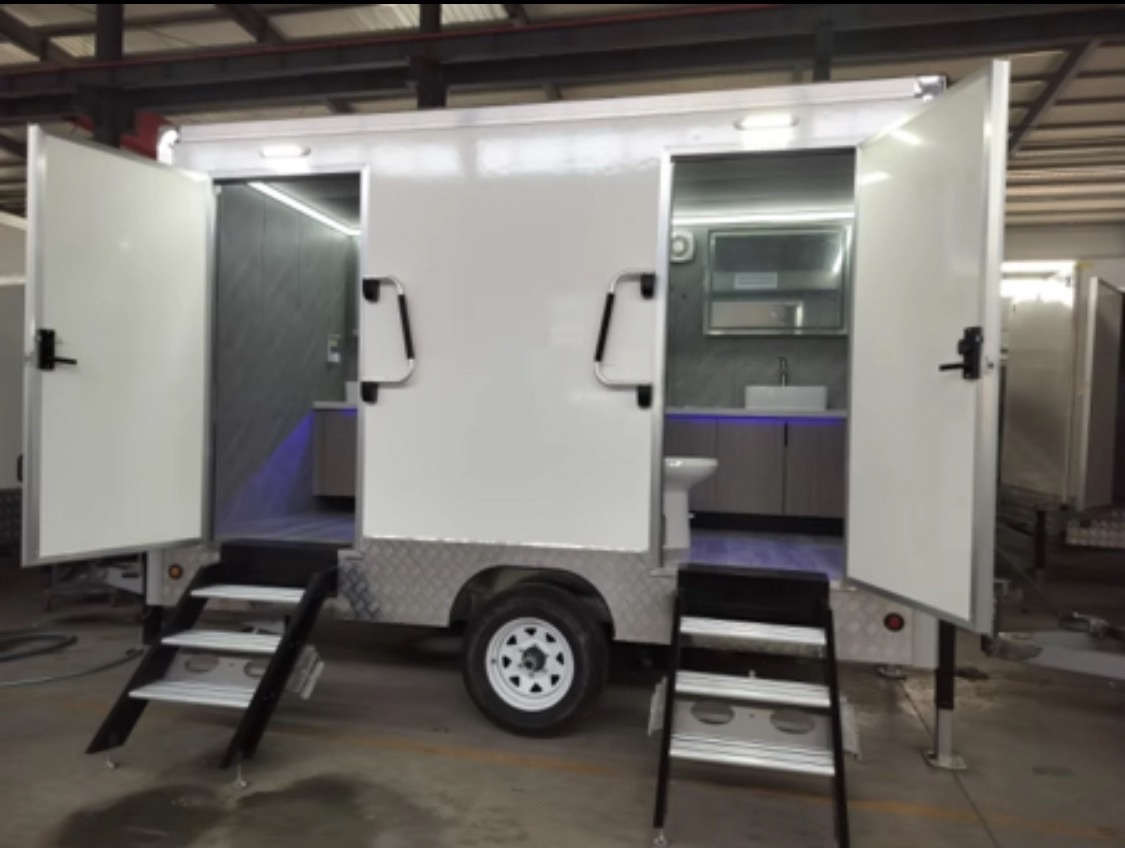 A mobile trailer with open doors revealing a small bathroom interior featuring a toilet and sink.