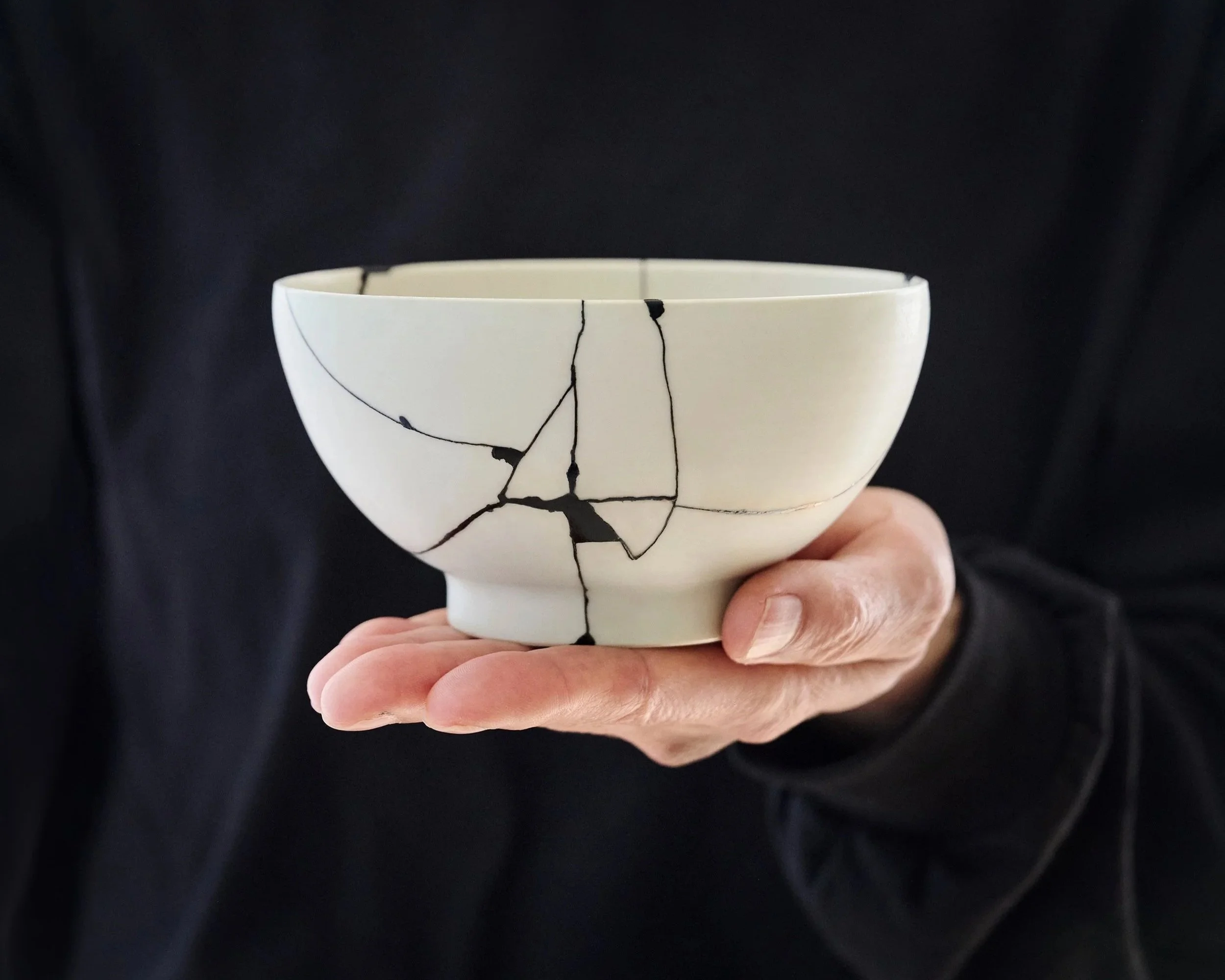 A hand holds a white ceramic bowl undergoing Kintsugi repair, with cracks painted in black Urushi as an intermediate stage.