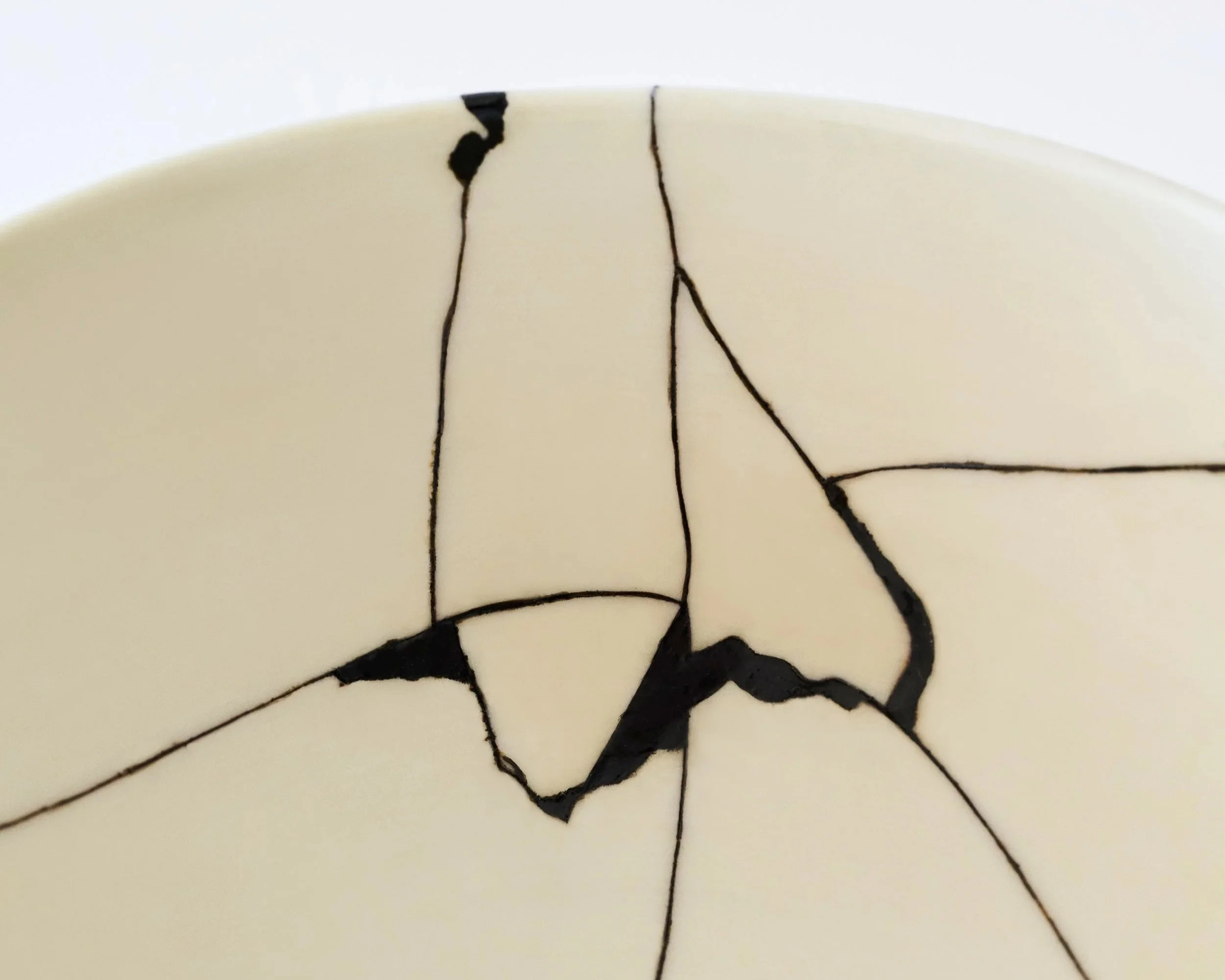 A close-up photograph shows a network of thin black lacquer lines mending the cracks of a repaired off-white ceramic bowl.
