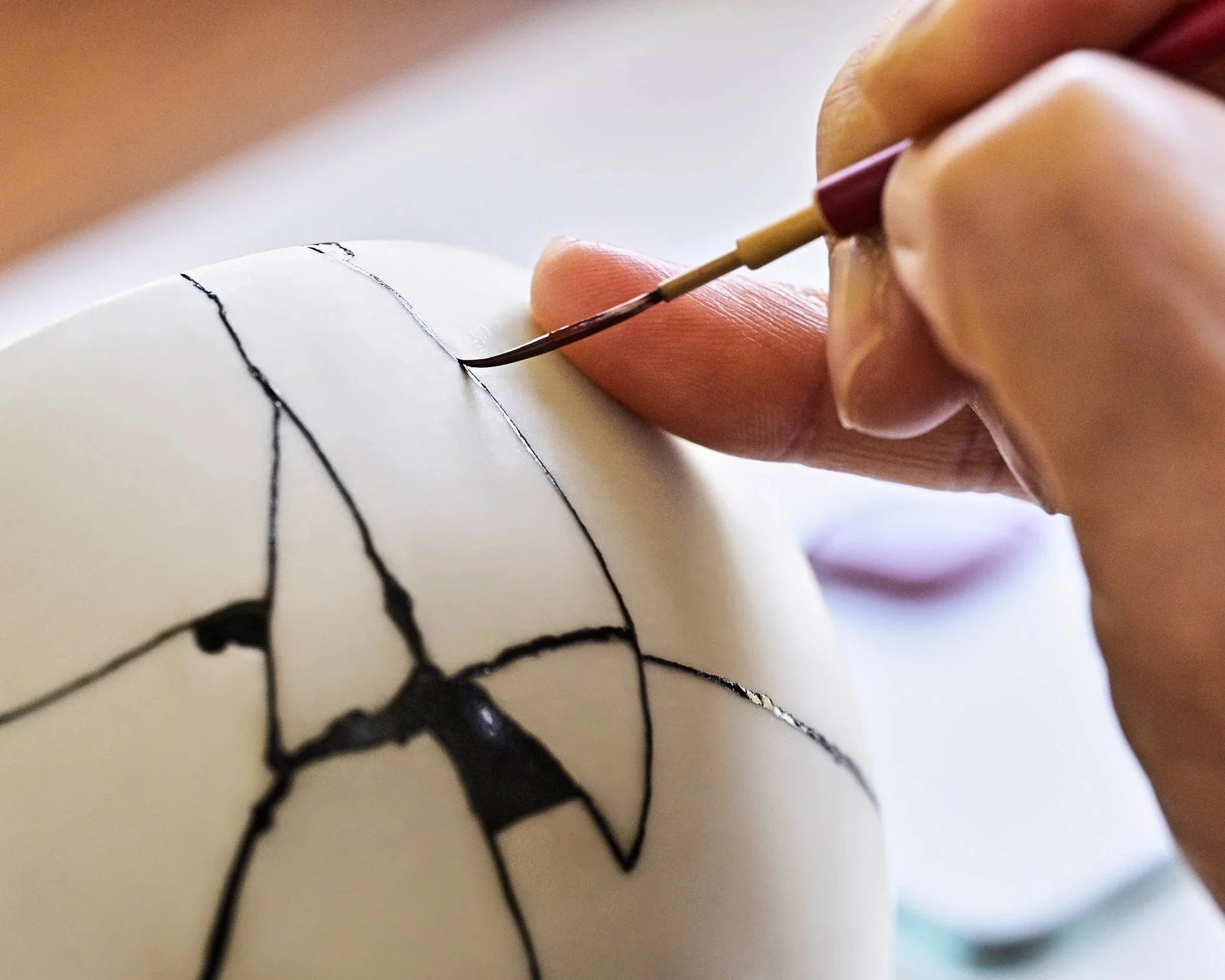 A close-up of a hand using a fine brush to carefully apply dark Bengala-urushi lacquer along the cracks of a white ceramic bowl.