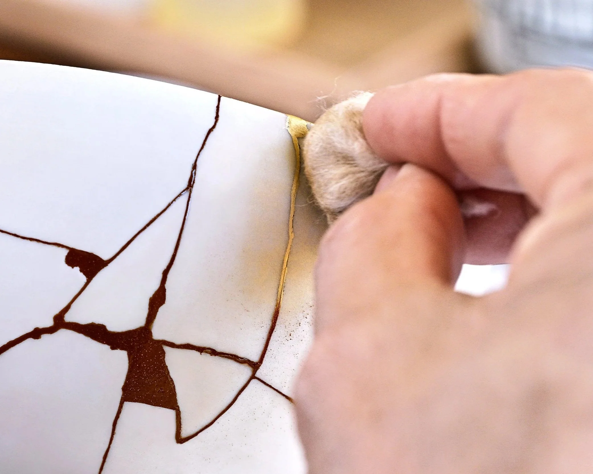 A close-up of a hand using a fine brush to carefully apply dark Bengala-urushi lacquer along the cracks of a white ceramic bowl.