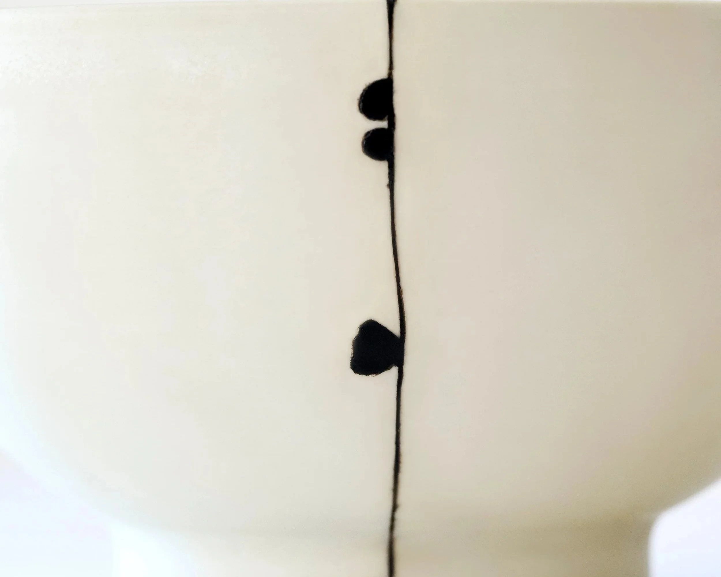 A close-up of a kintsugi repair in progress, with a thin line of black urushi lacquer and several graphic spots applied to a white ceramic surface.