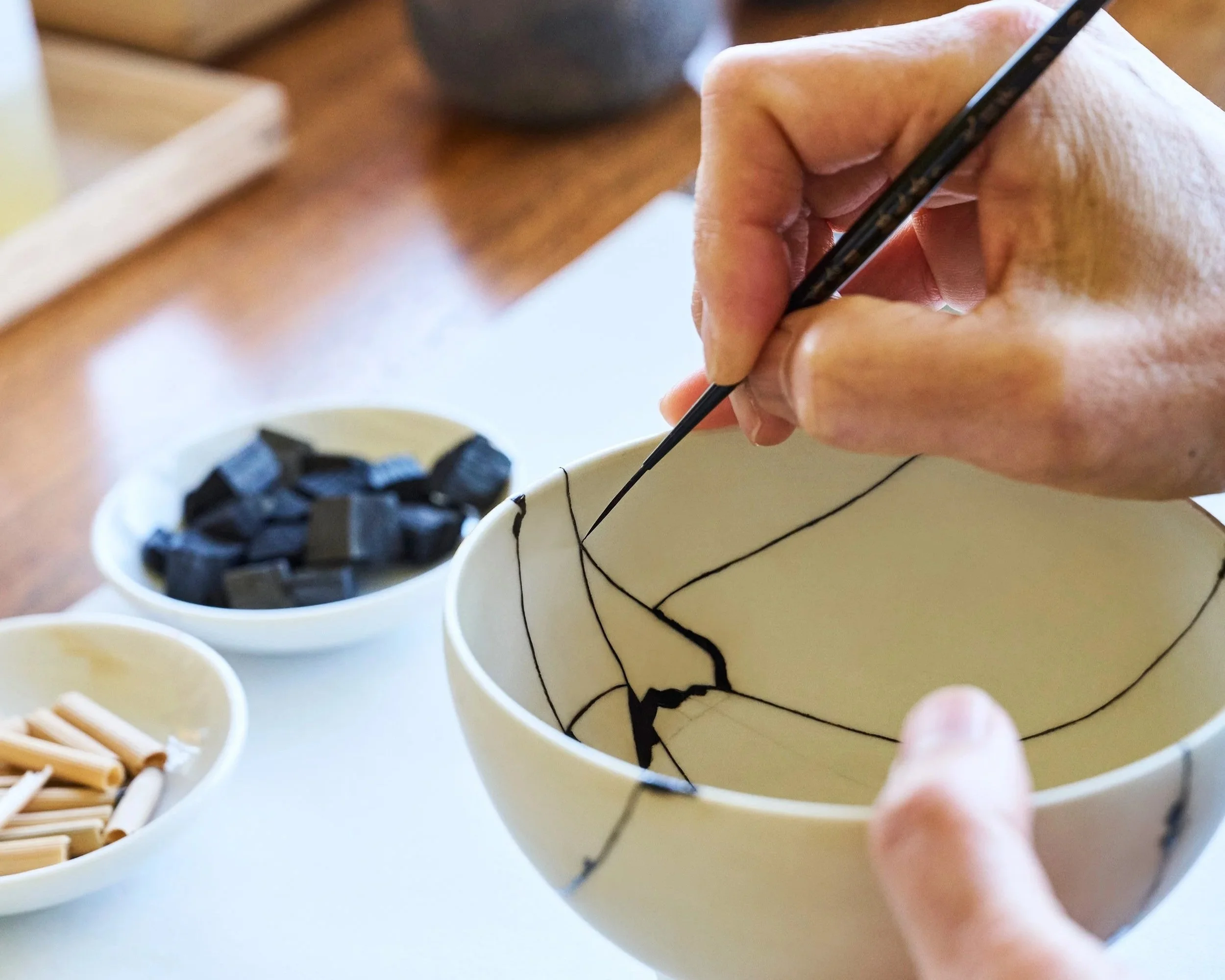 A close-up of a hand using a fine brush to carefully apply dark Bengala-urushi lacquer along the cracks of a white ceramic bowl.