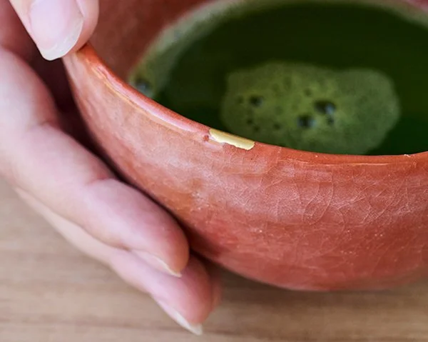 A hand holds a textured red-orange tea bowl filled with frothy green matcha, showcasing a delicate gold Kintsugi repair along the rim.