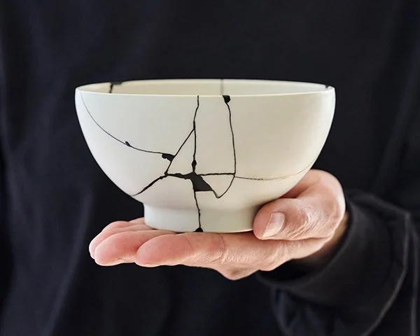 A hand holds a white ceramic bowl undergoing Kintsugi repair, with cracks painted in black Urushi as an intermediate stage.