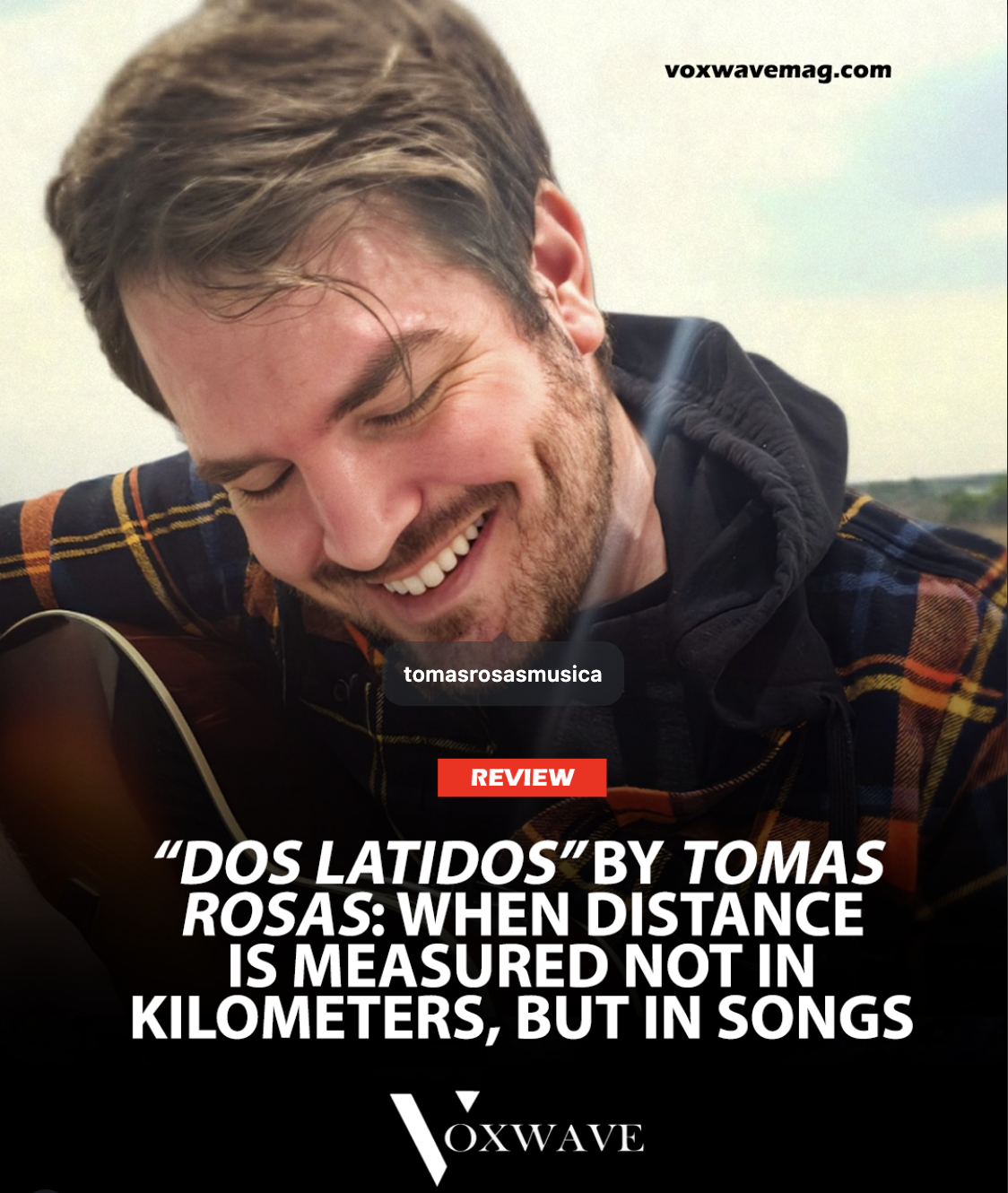 Close-up of a smiling man with closed eyes, playing an acoustic guitar outdoors on a sunny day, wearing a black hoodie and a plaid shirt.