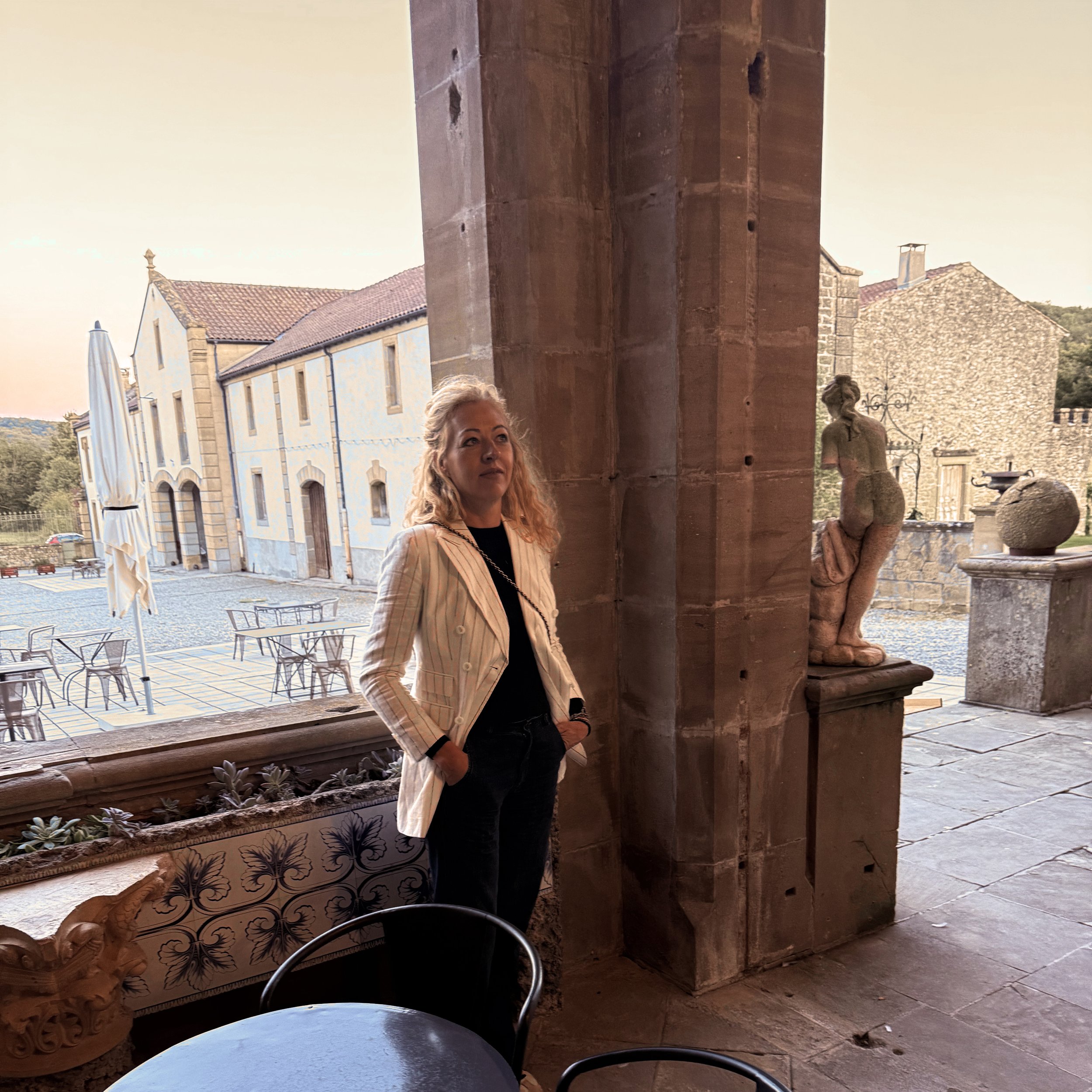 BIA LUCINA, Bronwyn Harrison A woman with blonde curly hair wearing a white striped blazer standing near a stone column inside a building with an outdoor courtyard in the background.