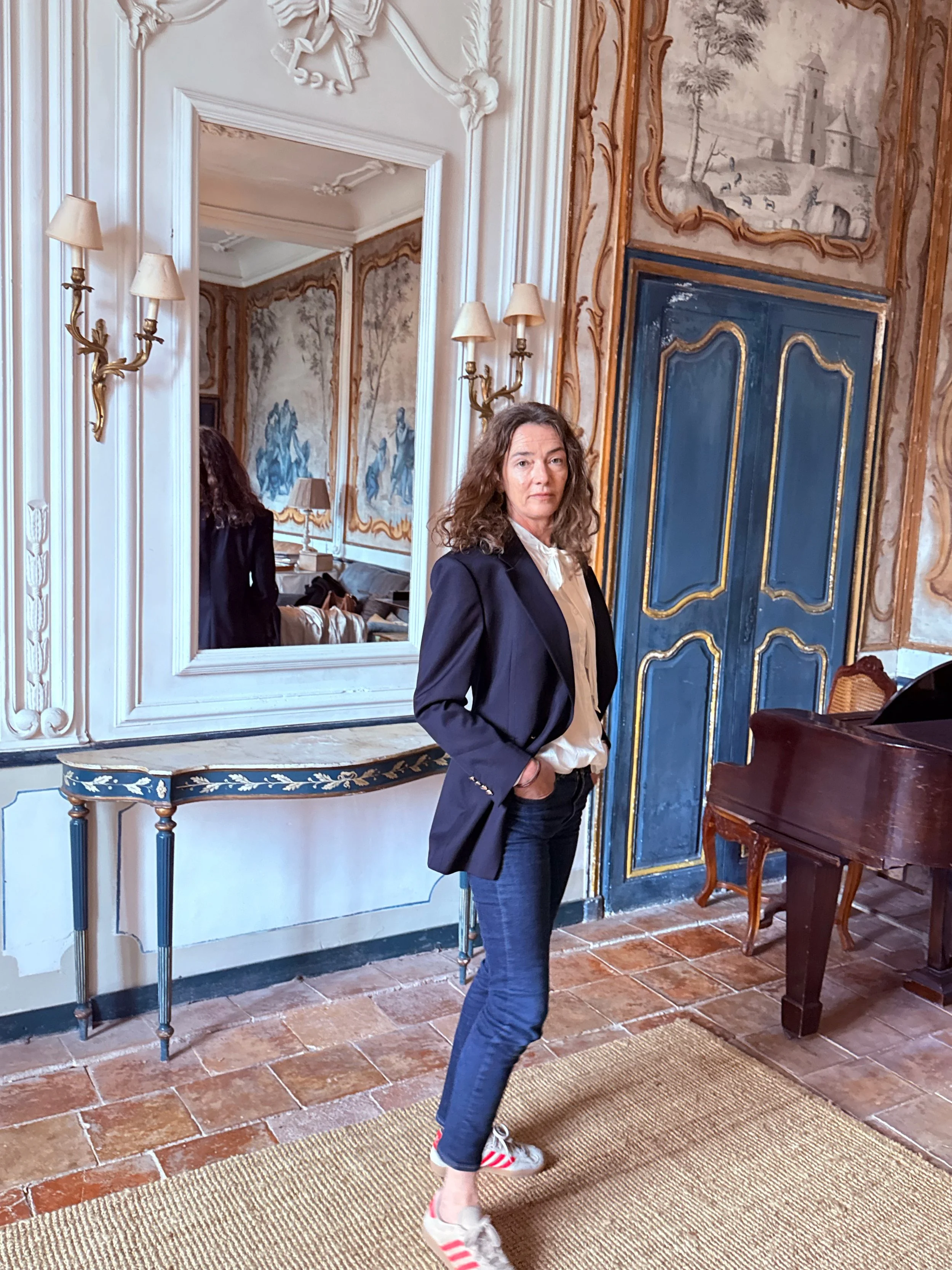 A woman with curly hair wearing a navy blazer, beige blouse, blue jeans, and sneakers with red and white stripes standing in an ornate room with decorative wall art, a large mirror, and a piano.