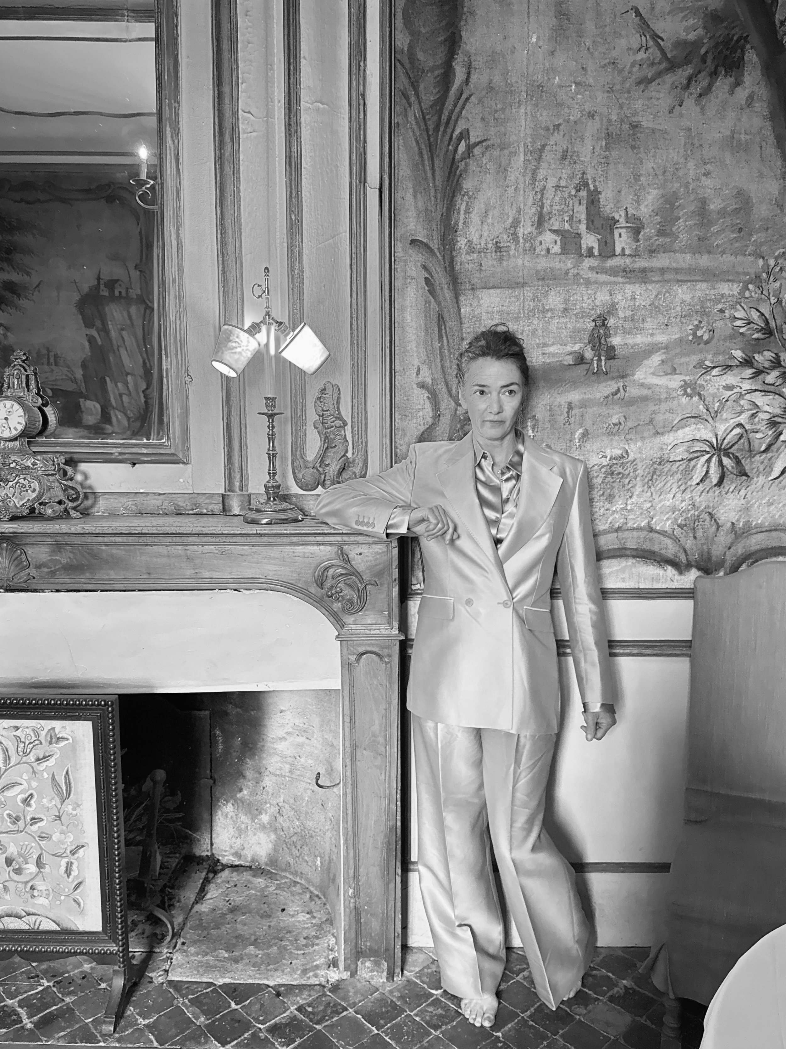 Black and white photo of a woman in a shiny satin suit standing in a room with vintage furniture and paintings, her right arm resting on a fireplace mantel, looking at the camera.