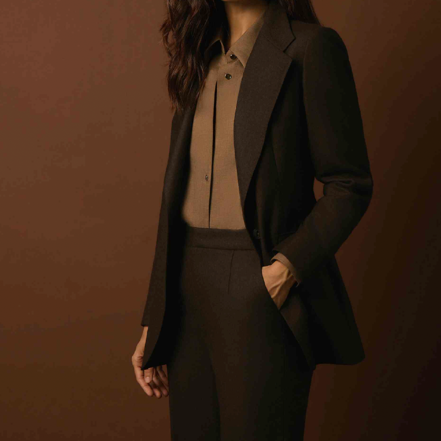 Women's Tailored custom made Woman in dark brown suit with beige shirt, standing against a brown background, with her left hand in her pocket.