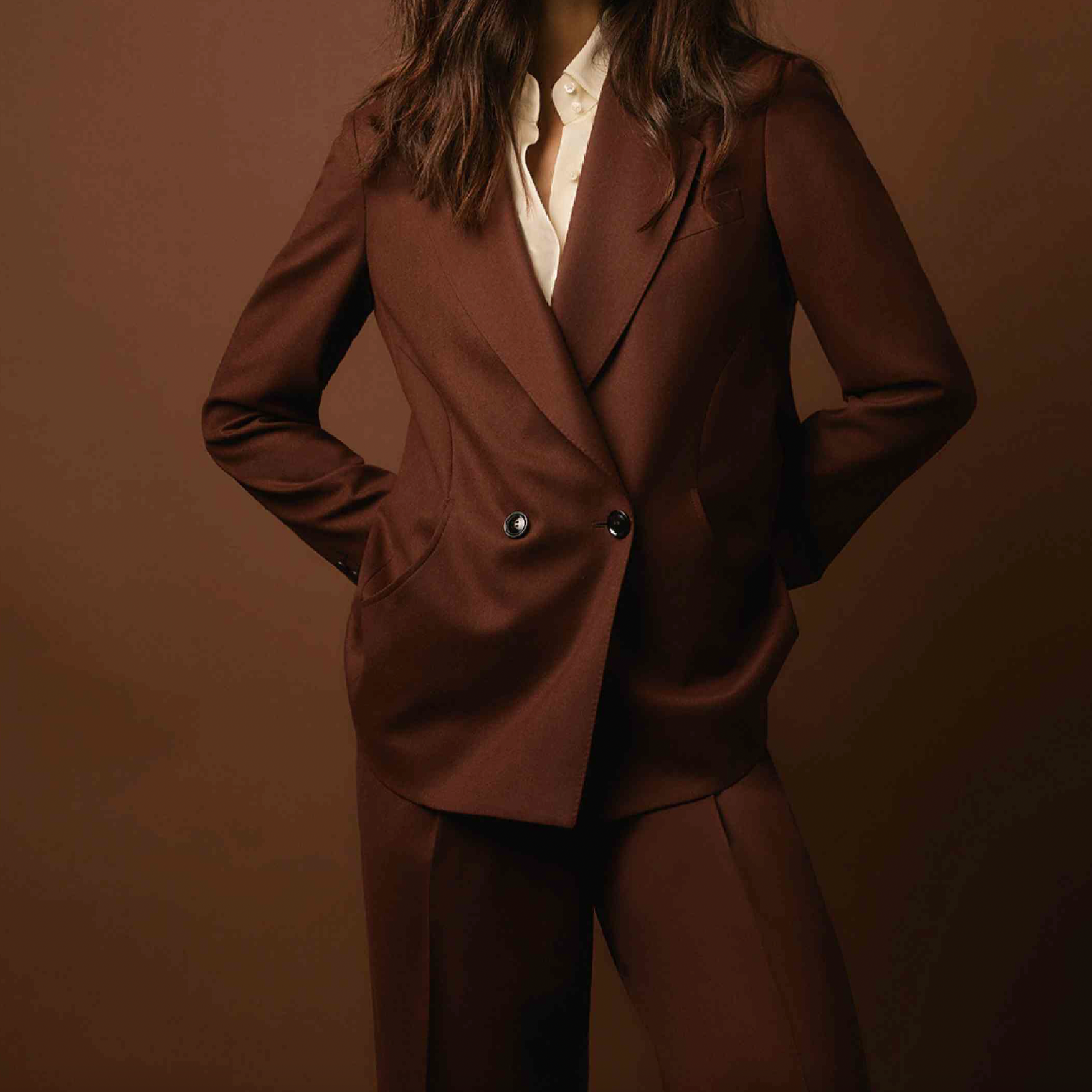 Women's Tailored custom made Woman in a brown suit with hands in pockets, standing against a brown background.