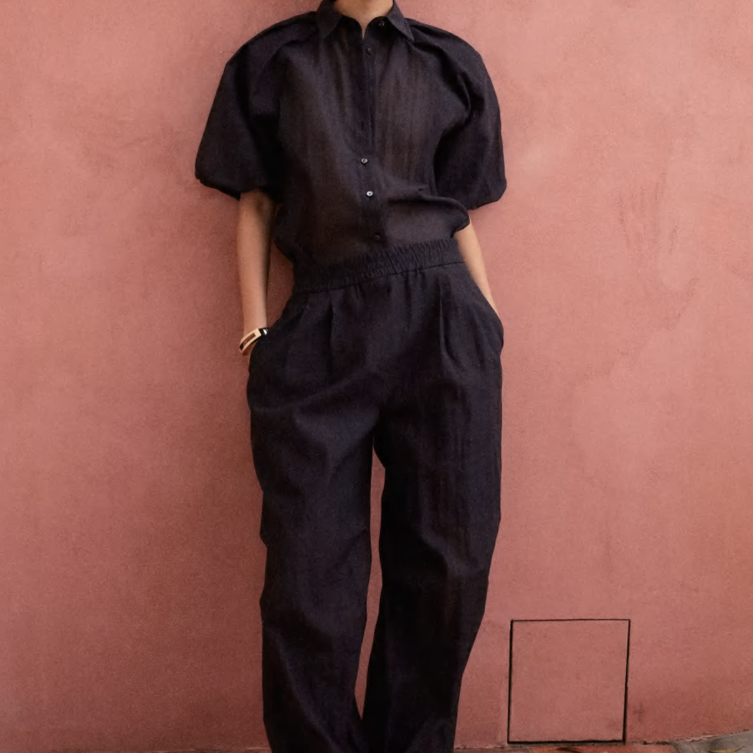 Women's Tailored custom made Person wearing a black short-sleeve shirt and matching black pants standing against a pink wall.