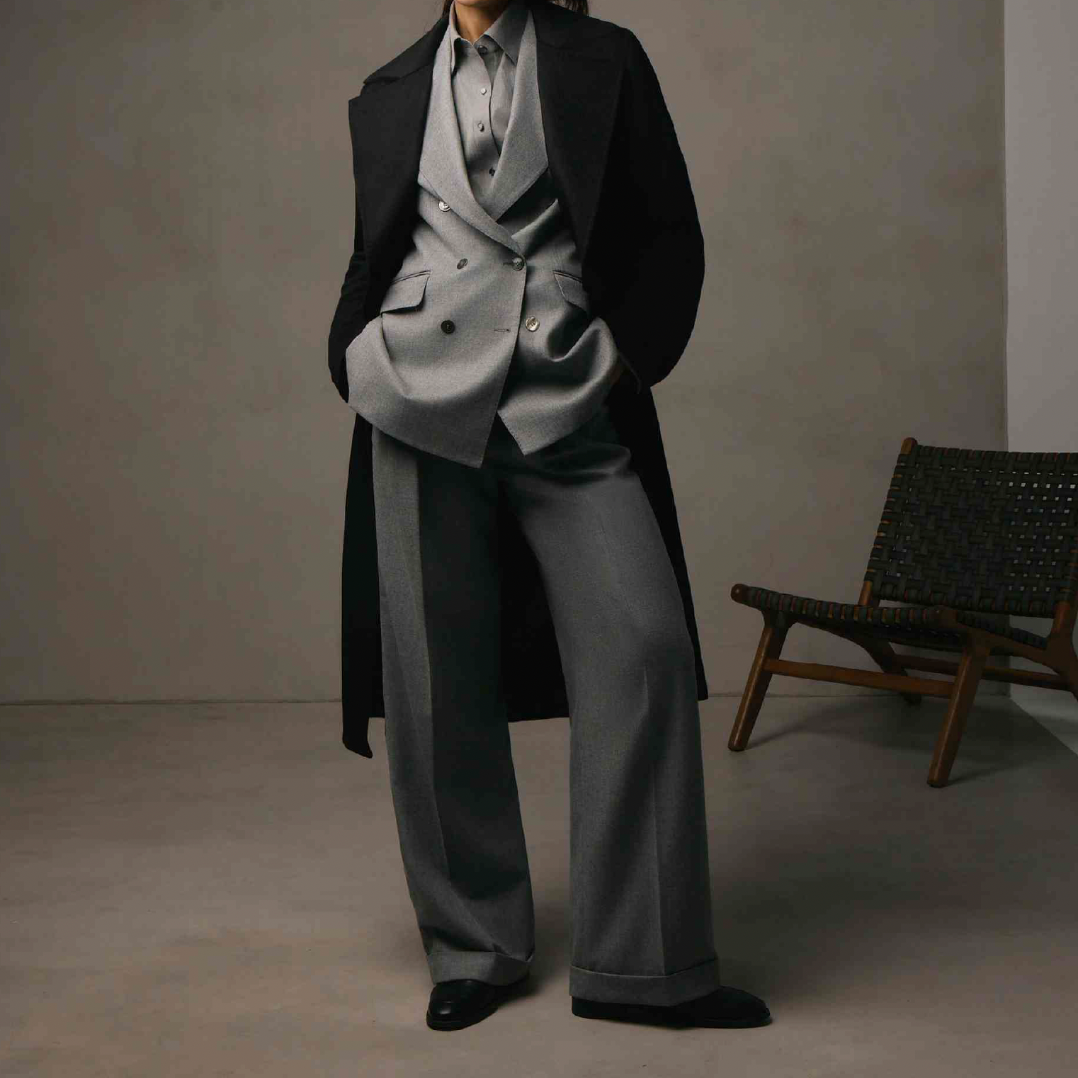 Women's Tailored custom made  Person wearing a gray suit with a black coat over it, standing next to a woven chair in a minimalist room.