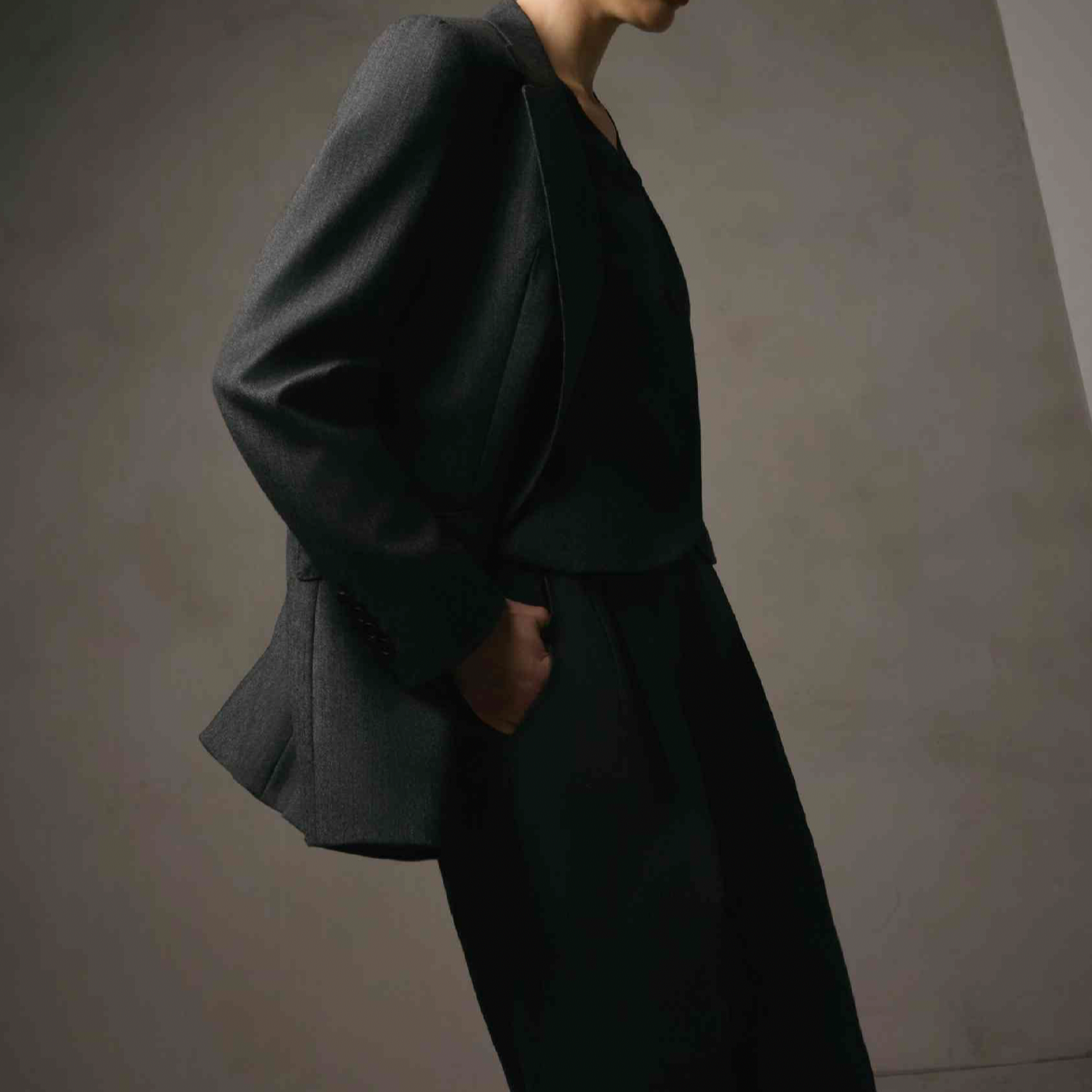 Women's Tailored custom made Person wearing a dark blazer and matching pants, with one hand in pocket, standing against a plain wall.