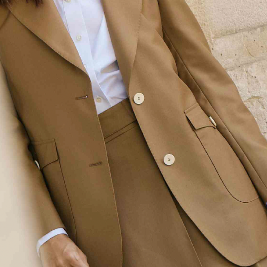 Women's Tailored custom made   Close-up of a person wearing a tan suit jacket and matching pants with a white dress shirt, against a beige wall.