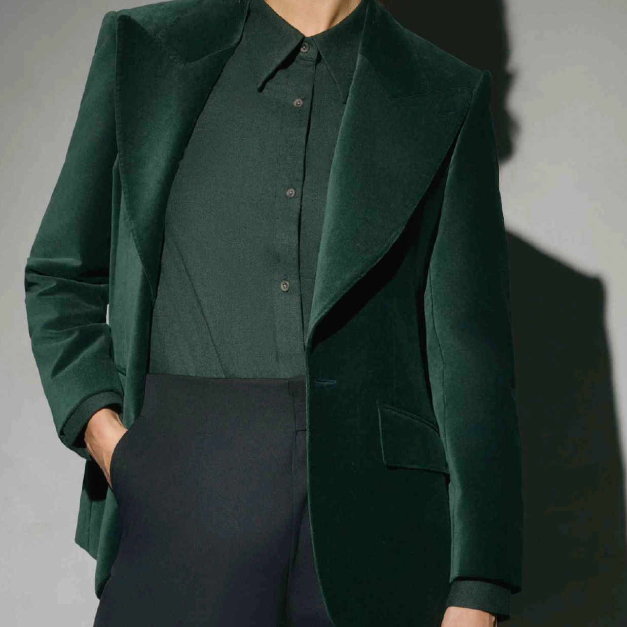 Women's Tailored custom made Person wearing a dark green blazer over a matching dark green button-up shirt and dark pants, with one hand in their pocket.