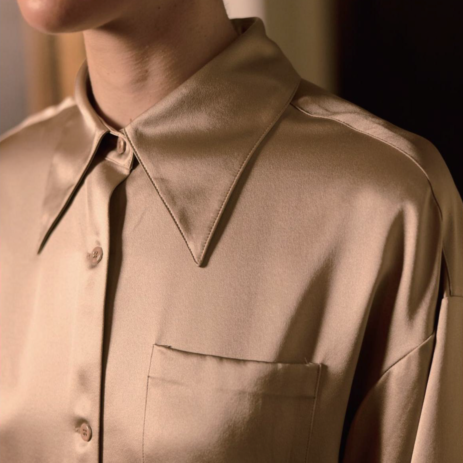 BIA LUCINA. womenswear Close-up of a beige satin shirt with a pointed collar and front buttons, part of a man's upper body.