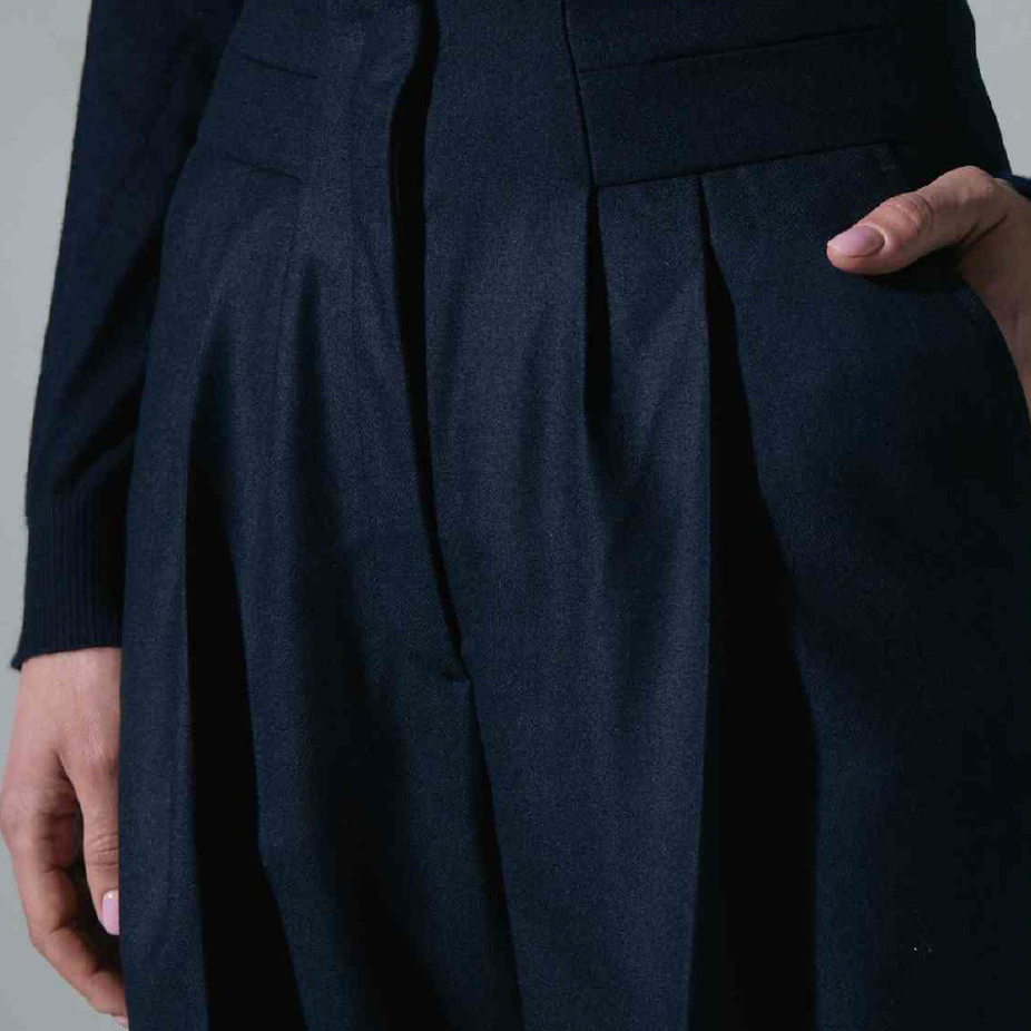 Women's Tailored custom made Close-up of a person's waist and legs wearing dark tailored trousers with pleats, a matching dark blazer, and a person's hand placed in the pocket.