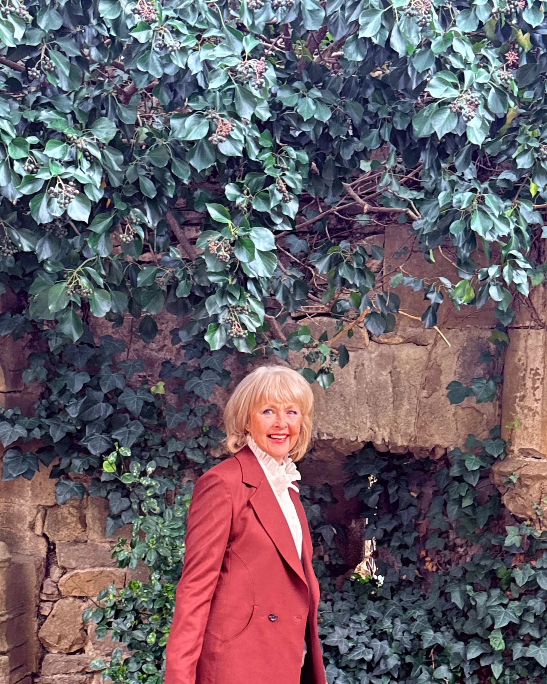 A woman with short blond hair smiling, wearing a rust-colored blazer over a white high-neck blouse, standing in front of a stone wall covered with green ivy.