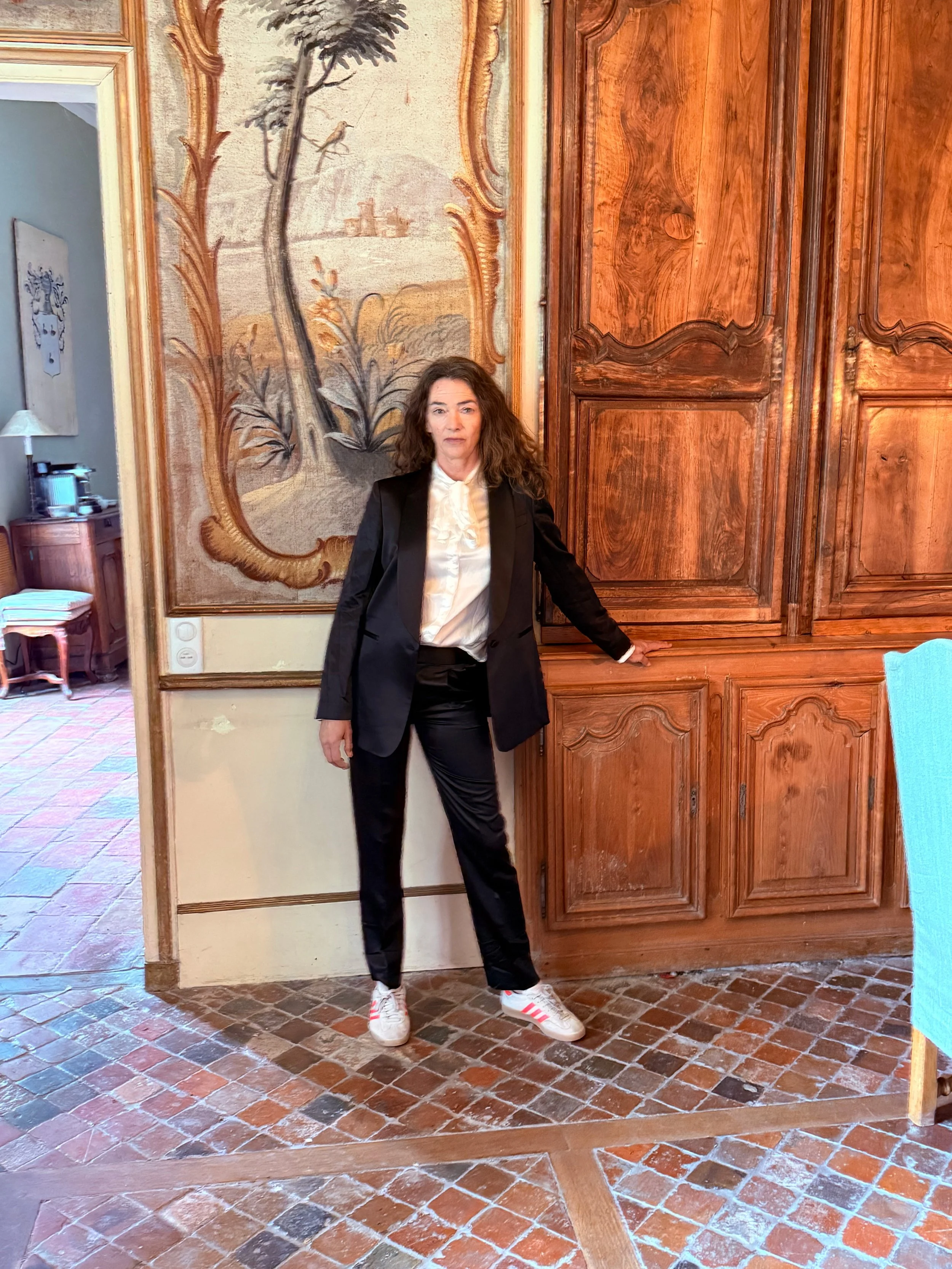 Woman with curly hair wearing a black suit with white shirt and sneakers, standing in a room with wooden furniture and decorative wall art.