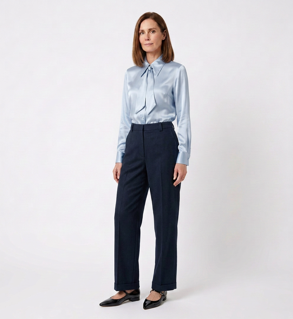 A woman with shoulder-length brown hair wearing a light blue satin blouse with a bow tie at the collar, dark trousers, and black shoes, standing against a plain white background.