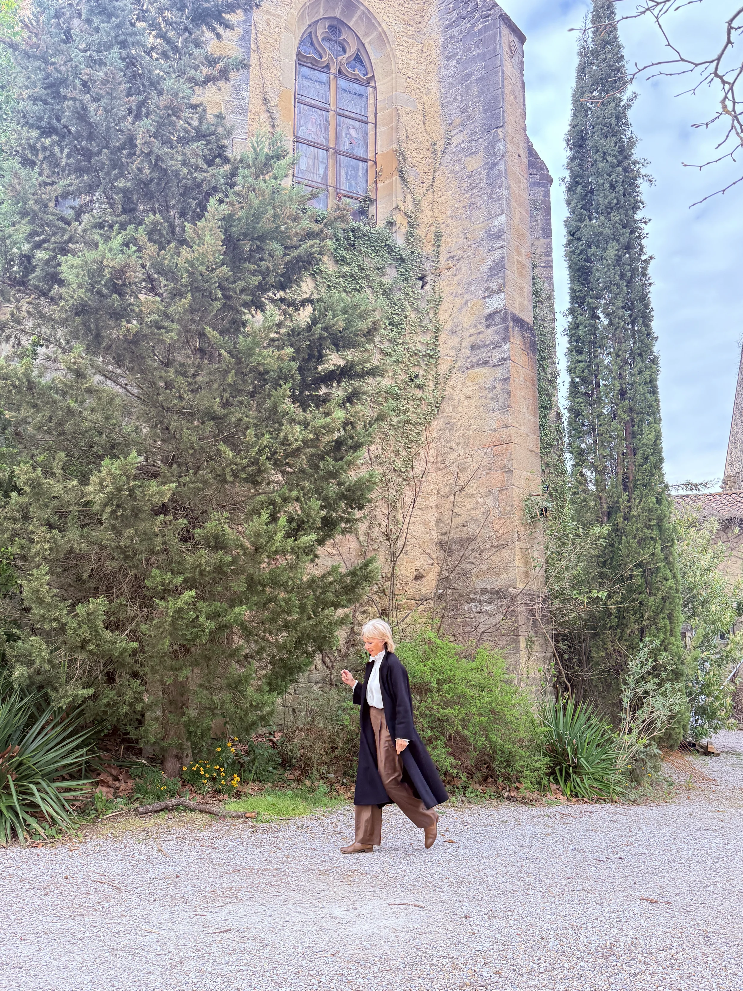 A woman with short gray hair, wearing a white shirt, black blazer, and brown wide-leg pants, walks along a gravel path in a garden with lush greenery. In the background, there is an old stone building with a large stained glass window and ivy growing on the wall. Tall cypress trees stand to the right of the building.