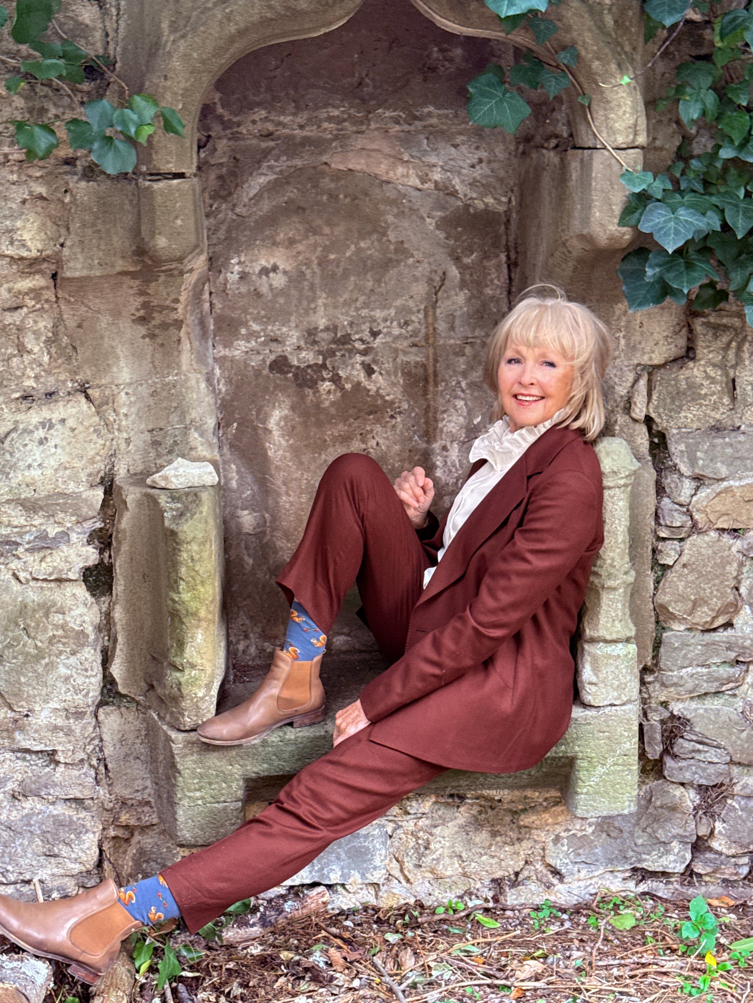 A smiling woman with blonde hair, wearing a brown suit and white shirt, sitting on a stone ledge in front of a stone wall with ivy leaves.