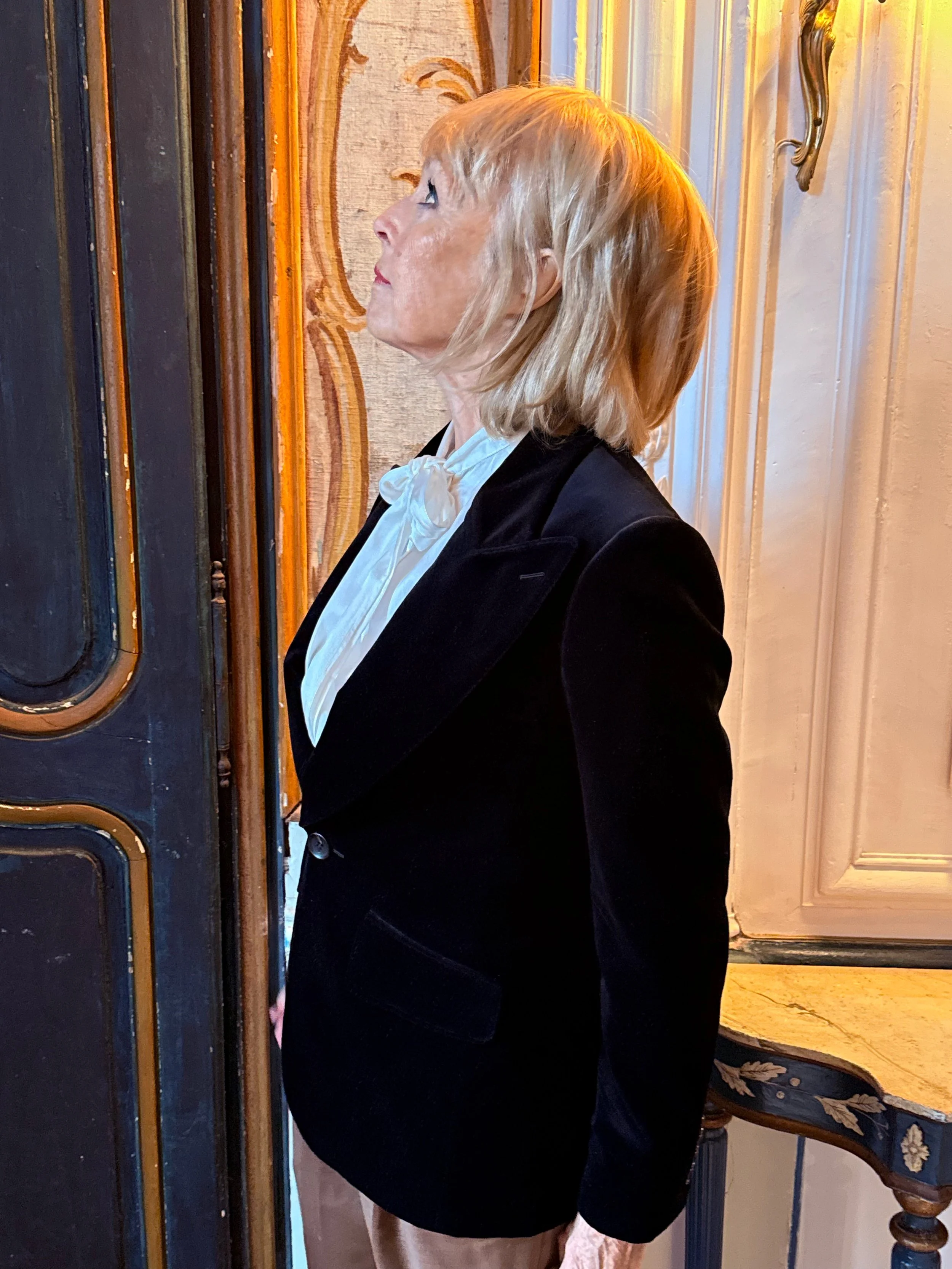 A profile of a middle-aged woman with short blonde hair, wearing a black blazer and a white blouse with a bow, standing indoors near a wooden door and decorative wall paneling.