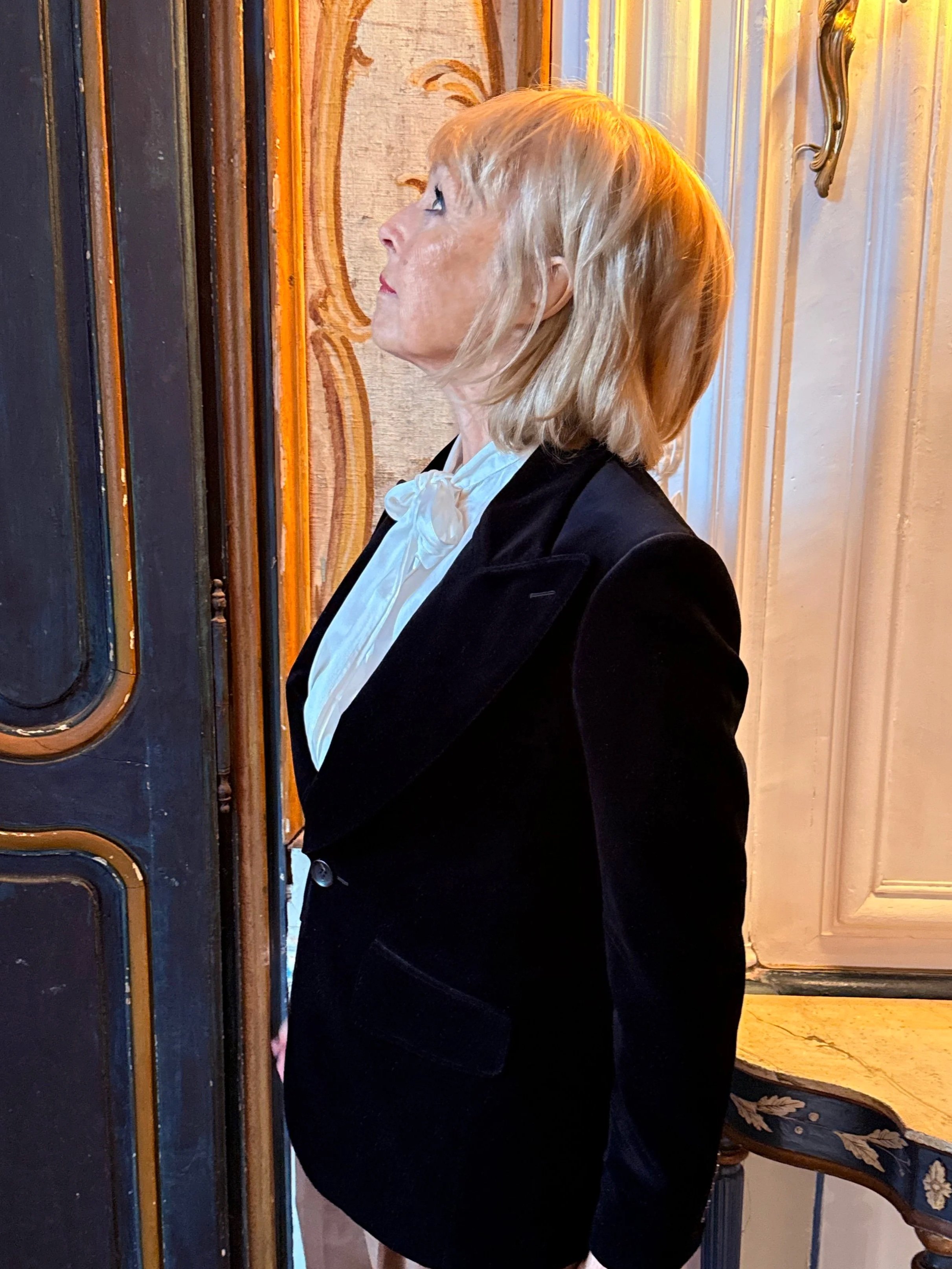 A woman with shoulder-length blonde hair, wearing a black jacket and white blouse with a bow, standing sideways near a patterned door with gold accents and a gold decorative element in the background.