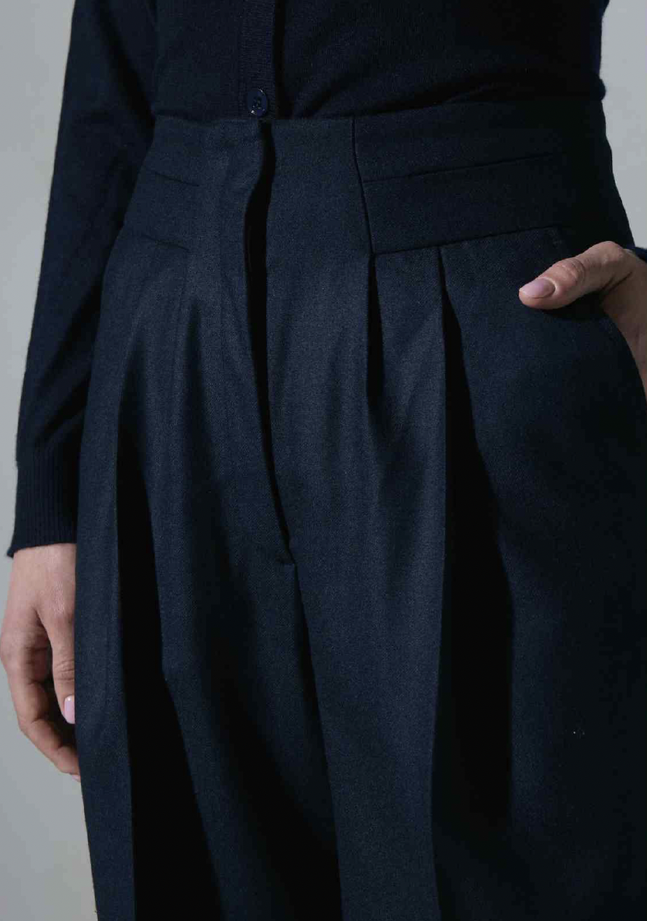 Women's Tailored custom made Close-up of a person's waist and legs wearing dark tailored trousers with pleats, a matching dark blazer, and a person's hand placed in the pocket.