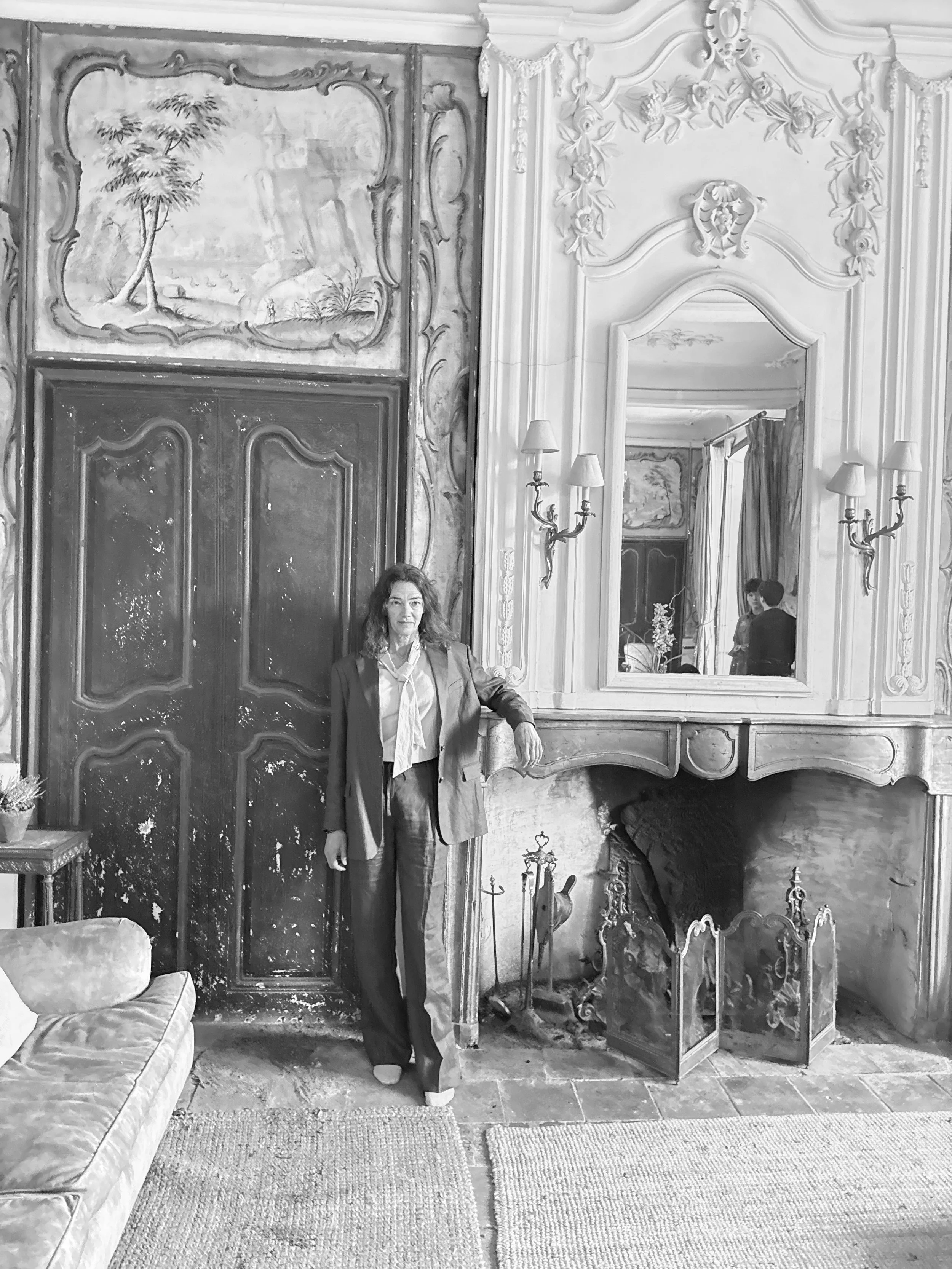 A woman dressed in a suit standing in an elegant, ornate room with elaborate wall decorations, a large mirror, and a fireplace.