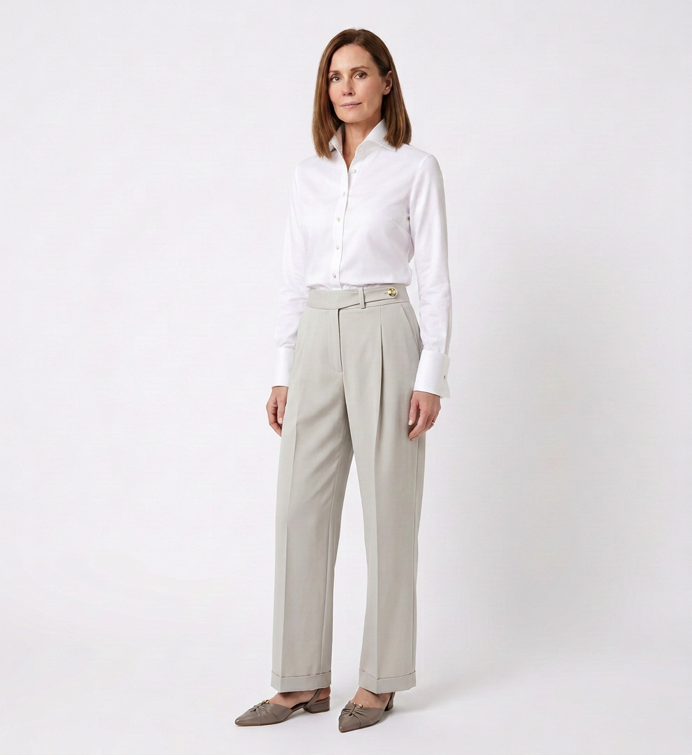 A woman with shoulder-length brown hair wearing a white button-up shirt tucked into high-waisted beige trousers and gray shoes, standing against a plain white background.