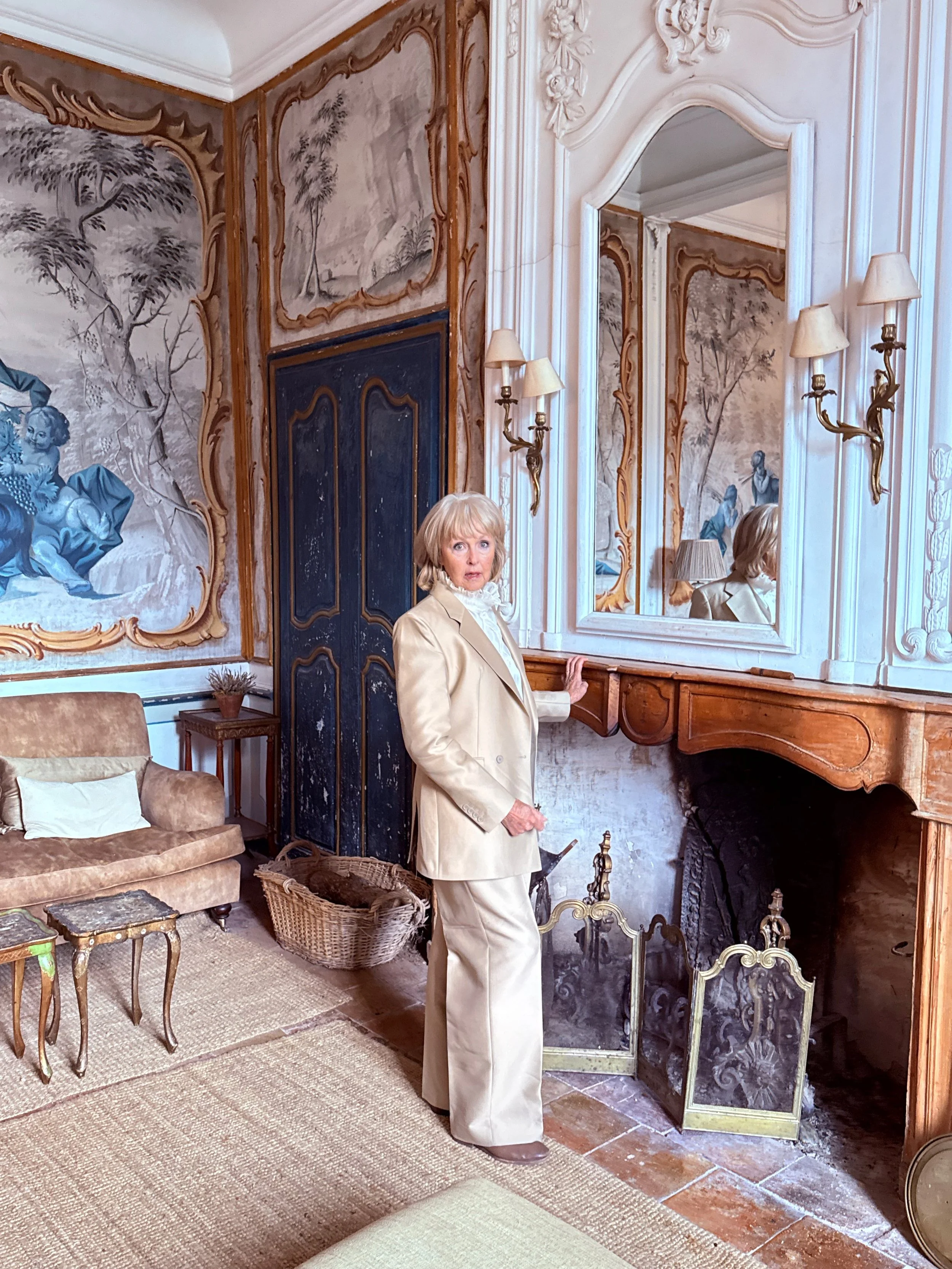 A woman standing in a richly decorated room with ornate wall murals, mirrors, and antique furniture. She is wearing a beige suit and looking at the camera.