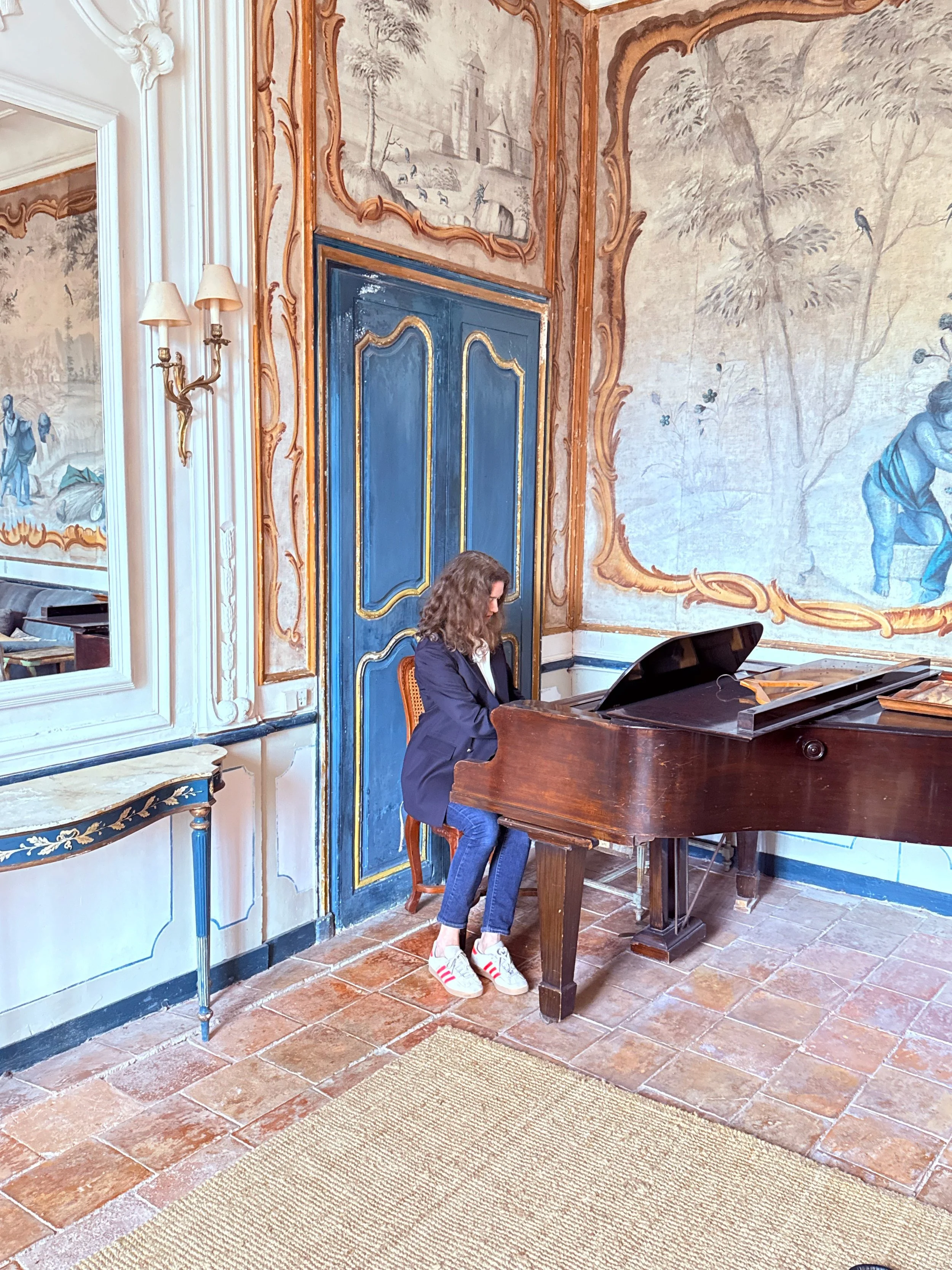 A woman playing a grand piano in an ornate room with decorative painted walls, a blue door, a mirror, and a wall sconce.